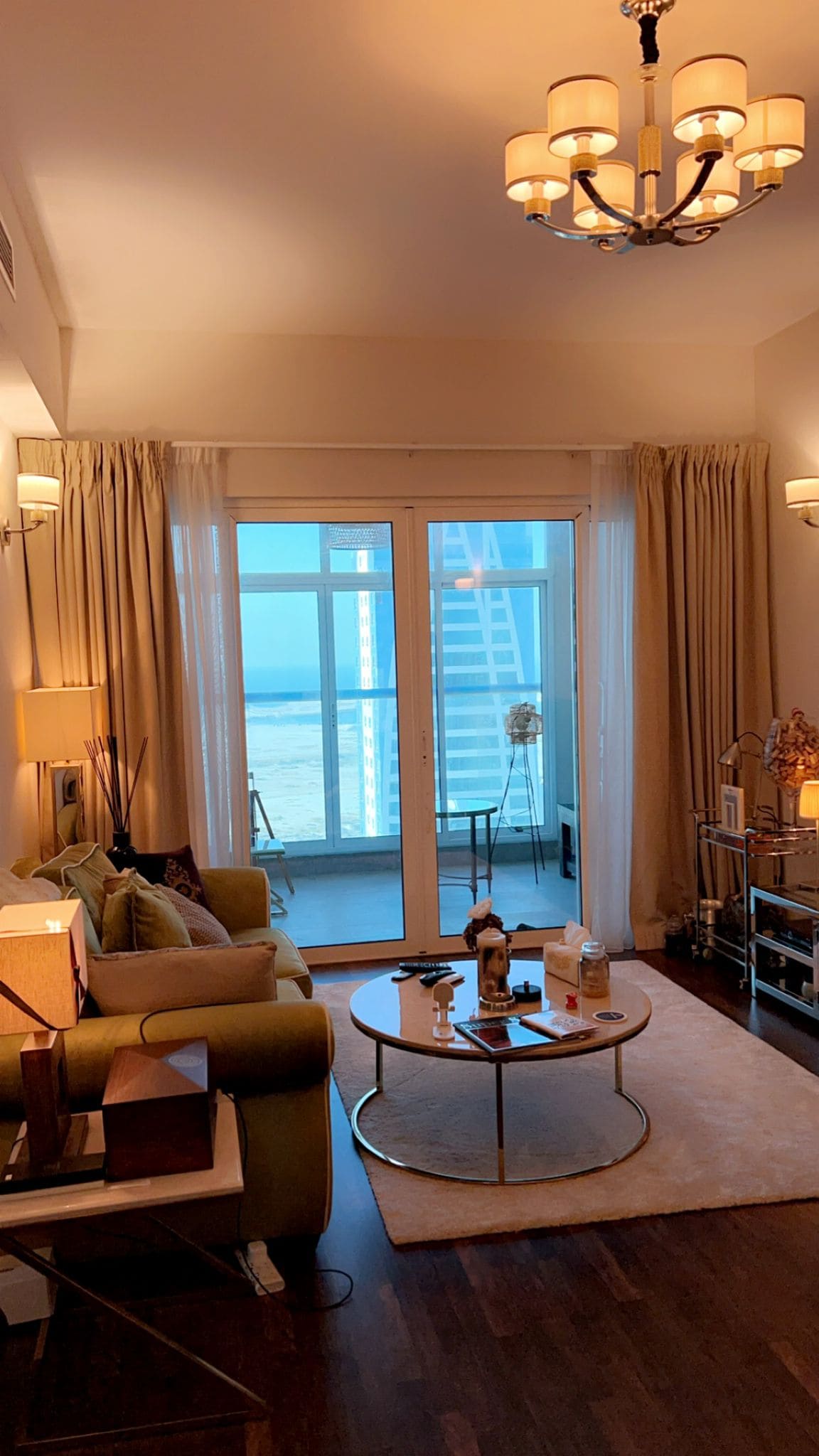 A living room with a green sofa, round coffee table, and chandelier. Large windows with curtains open to a balcony, offering a view of the sea. A side table and decor items are visible.