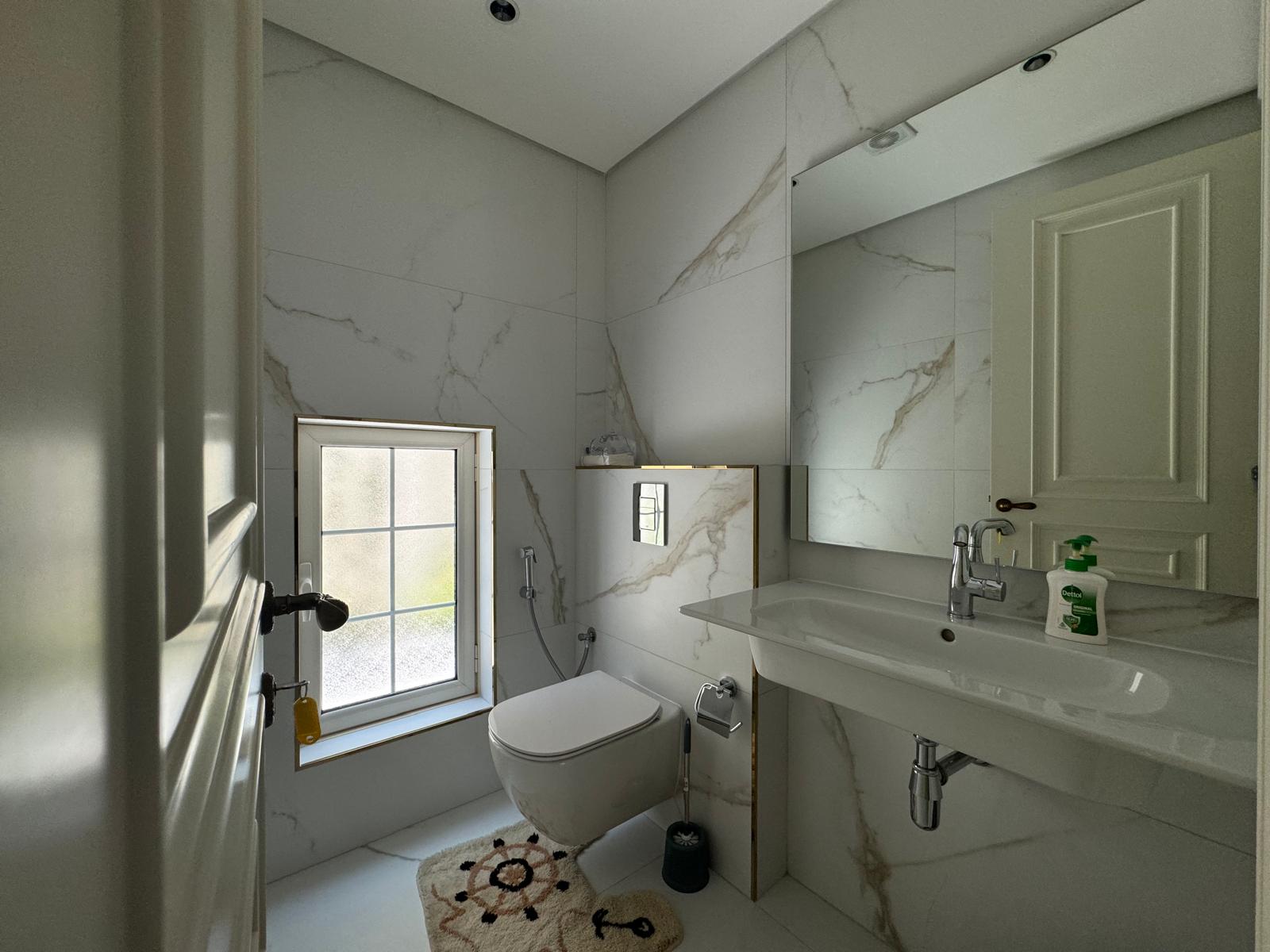 Modern bathroom with white marble walls, a wall-mounted toilet, sink with mirror, and a small window. Soap dispensers are on the sink.