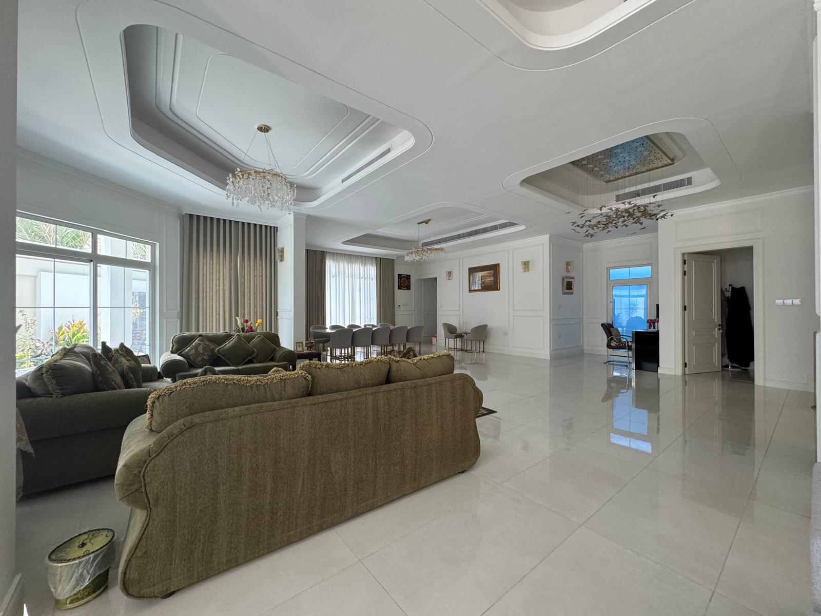 Spacious living room with green sofas, tile flooring, and a large dining table. Chandeliers hang from a coffered ceiling. Large windows allow natural light. Art decorates the white walls.