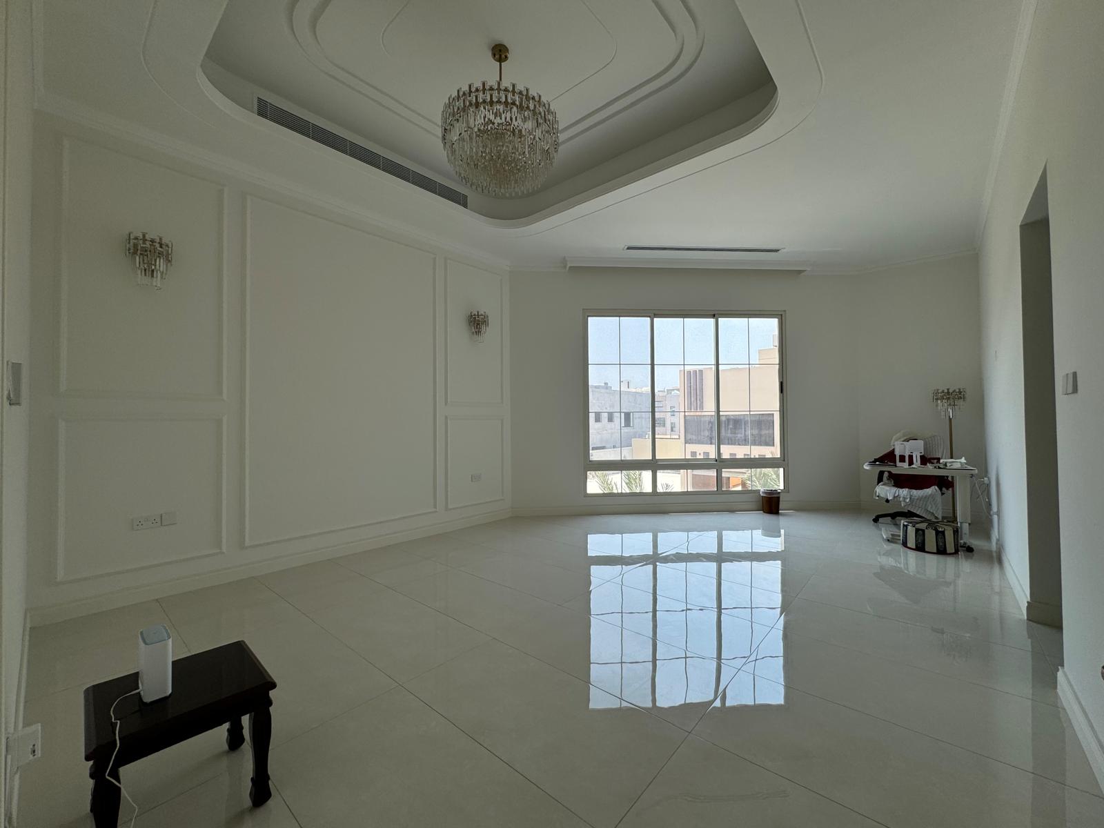 An empty, unfurnished room with white walls, glossy tiled floor, chandelier, and large window. A small table with items and bags is in the corner.