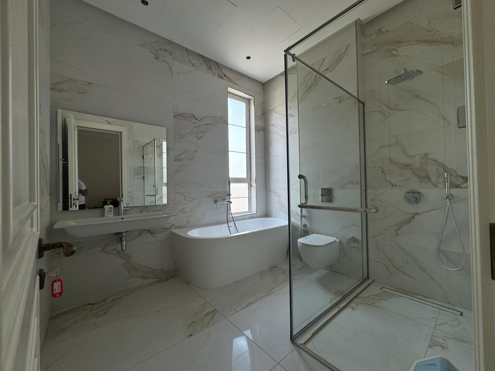 Modern bathroom with gray marble tiles, featuring a bathtub, glass-enclosed shower, toilet, sink with a mirror, and a window for natural light.