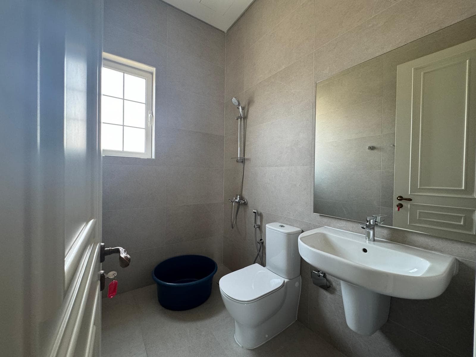 Bathroom with a sink, toilet, mirror, and wall-mounted showerhead. A small blue bucket is on the floor near the window. Walls are light grey tile.