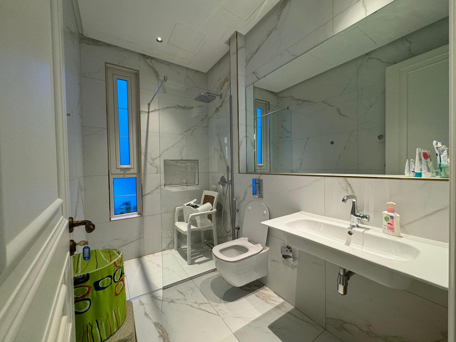 Modern bathroom with marble walls and floor. Features a glass shower enclosure, floating toilet, white sink, and a green wastebasket. Various toiletries are on the sink.