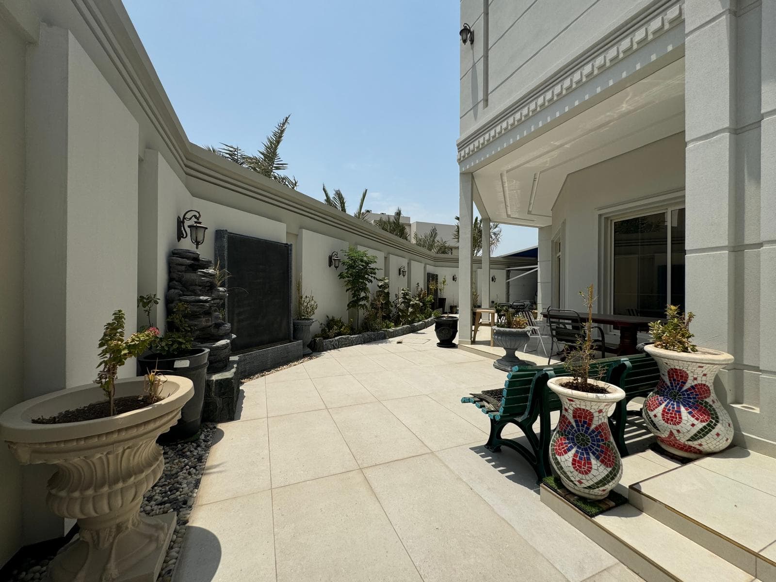 Sunny courtyard with white walls, potted plants, mosaic planters, and a small garden fountain feature. A green plastic chair sits by a table under a covered area.