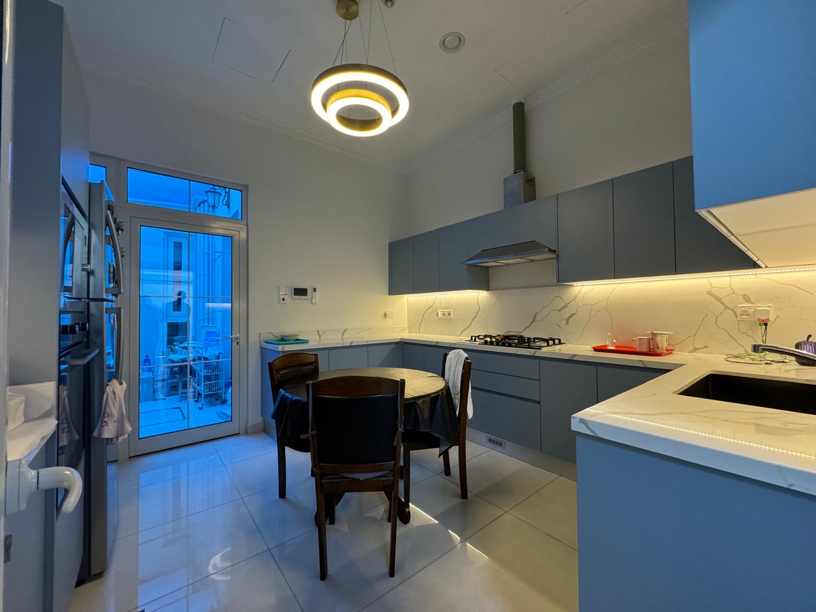 Modern kitchen with blue cabinets, built-in oven, and a round dining table with four chairs. A circular ceiling light is above the table. A small laundry area is visible through a glass door.