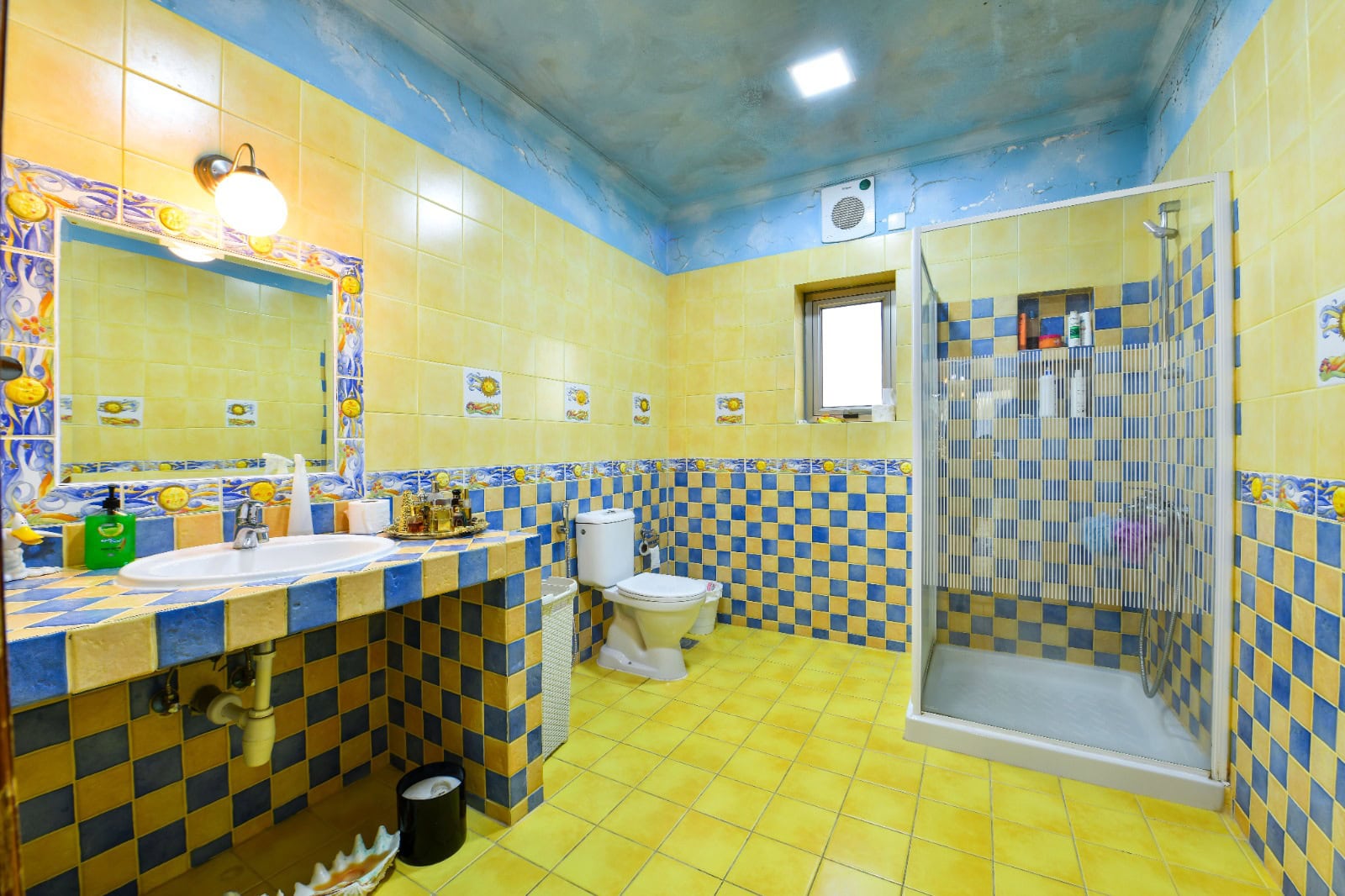 Bathroom with yellow and blue tiled walls and floor, a shower with glass doors, a toilet, a sink with a mirror above, and a ceiling-mounted light fixture.