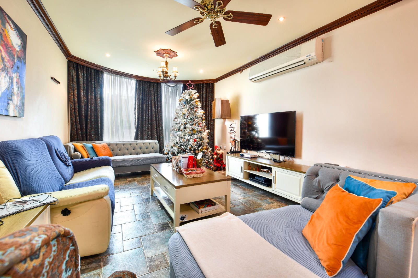 A cozy living room with a Christmas tree, couches with colorful pillows, a TV on a white stand, and a ceiling fan. The room has a wooden floor and a large window with dark curtains.