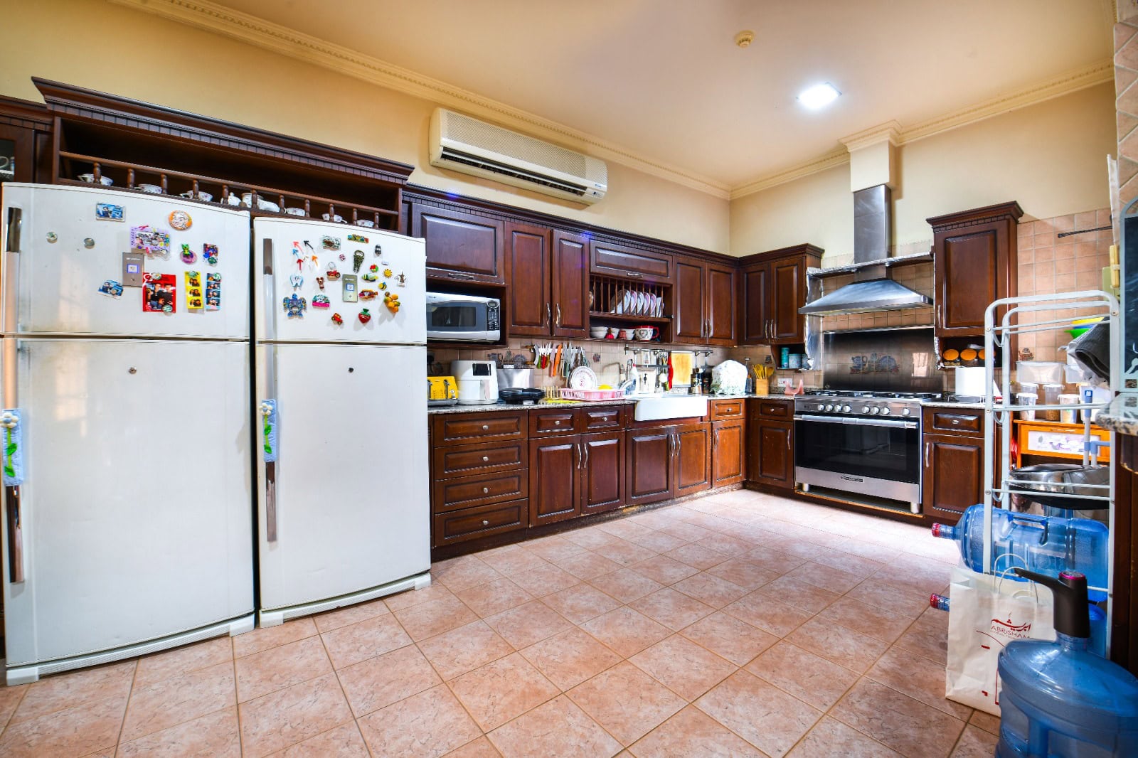 A spacious kitchen with dark wooden cabinets, two refrigerators with magnets, a large stove, microwave, and various kitchen items. The floor is tiled, and there are water dispensers on the side.