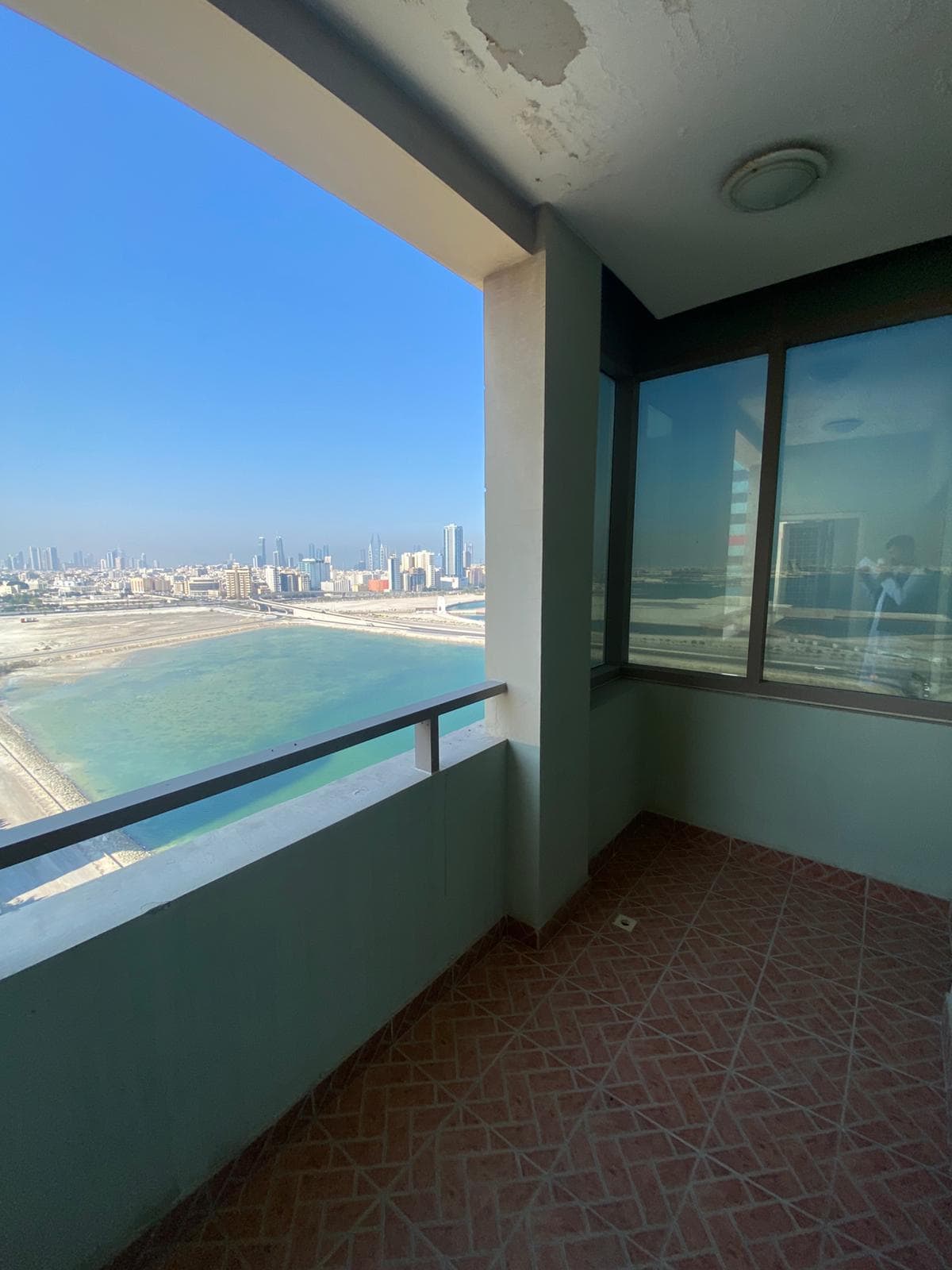 Balcony view of a cityscape with tall buildings, overlooking a body of water and shoreline. The balcony has tiled flooring and is partially enclosed with a railing and walls.