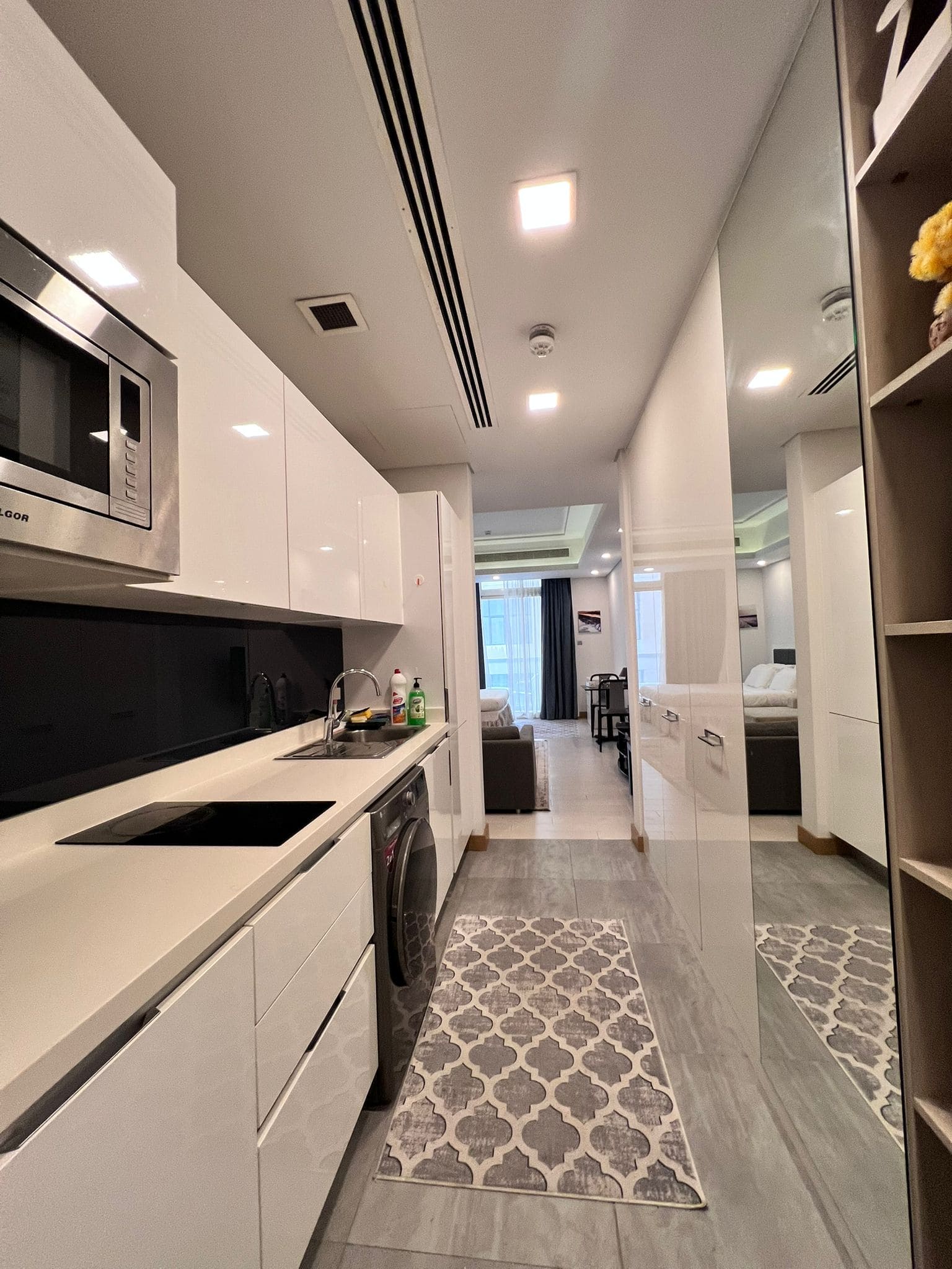 Modern apartment interior with a compact kitchen featuring white cabinets, a microwave, and an induction cooktop. The living area with a sofa is visible in the background.