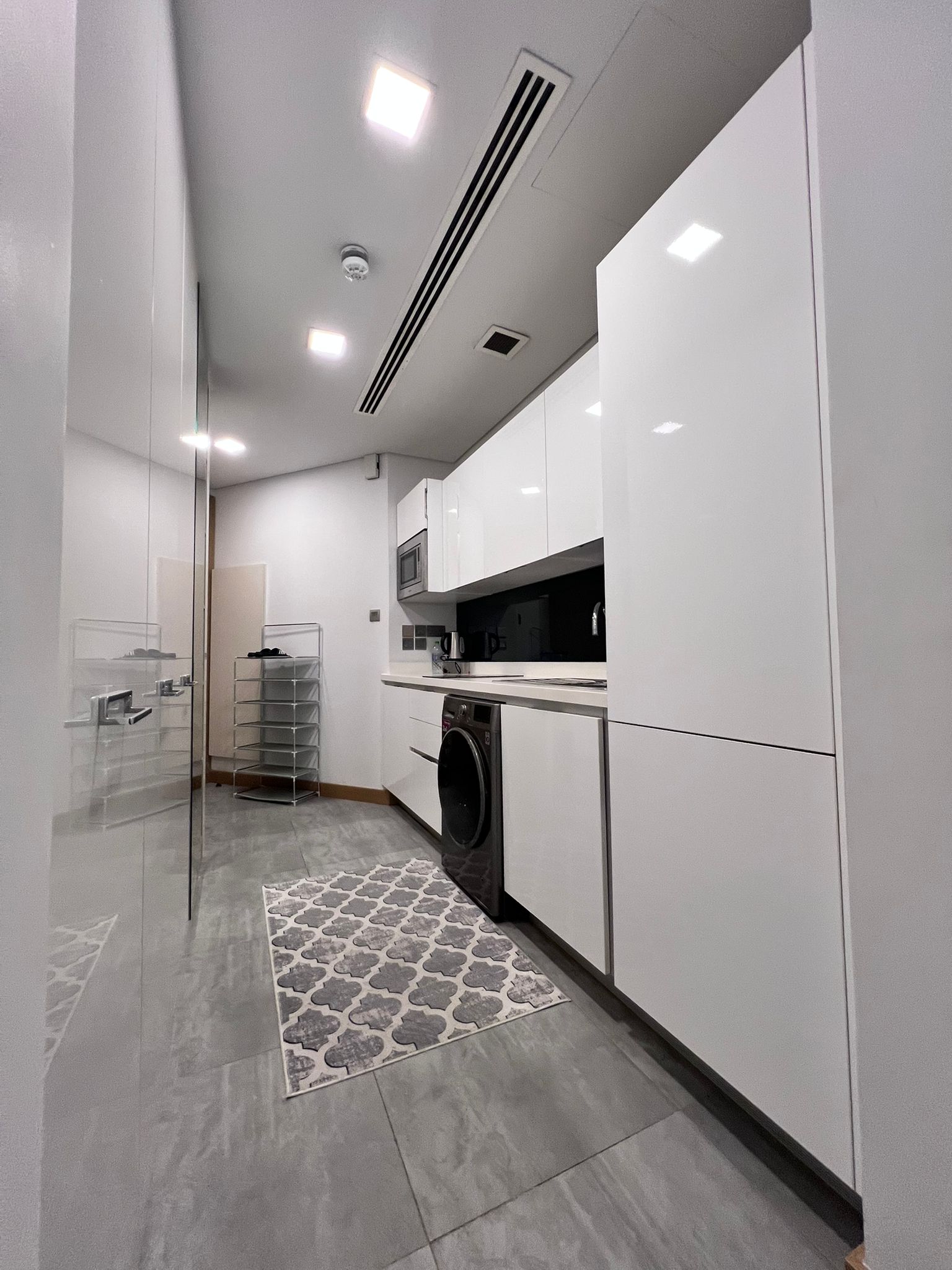 Modern kitchen with glossy white cabinets, a washer, microwave, and a patterned rug on the gray tile floor. Ceiling lights illuminate the space.