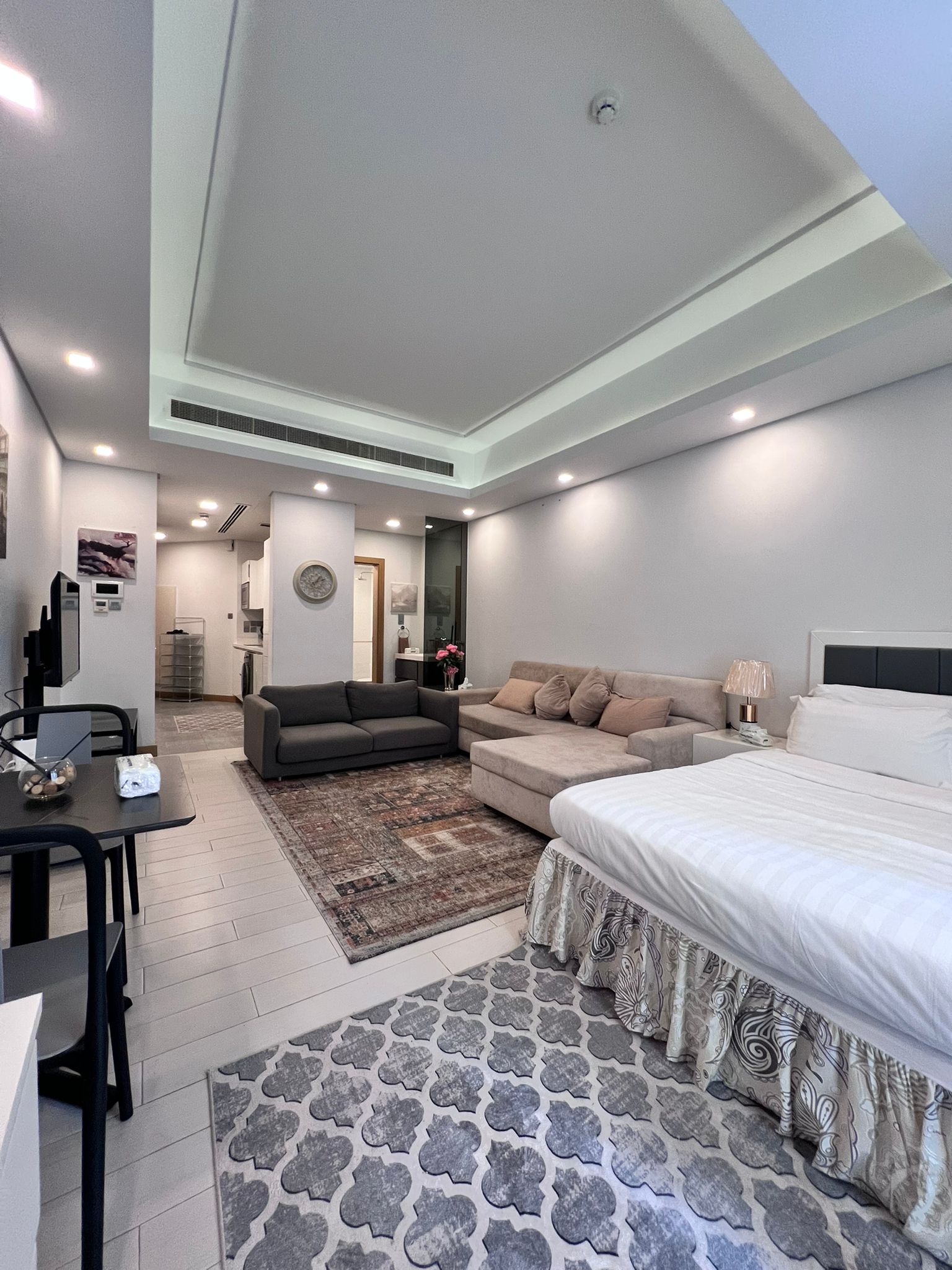 Modern studio apartment with a bed, gray sofa, and dining table. The room features a patterned rug, neutral tones, and recessed lighting. The kitchen is visible in the background.