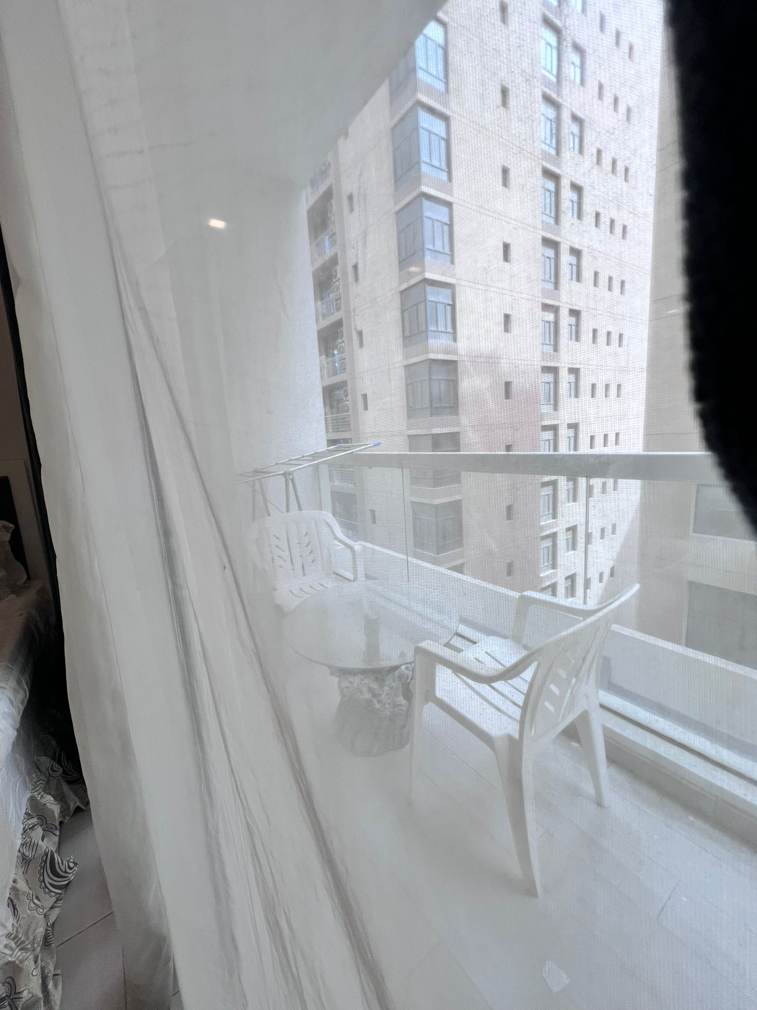 View of a high-rise building from a balcony with a glass railing, featuring white plastic chairs and a table behind a sheer curtain.