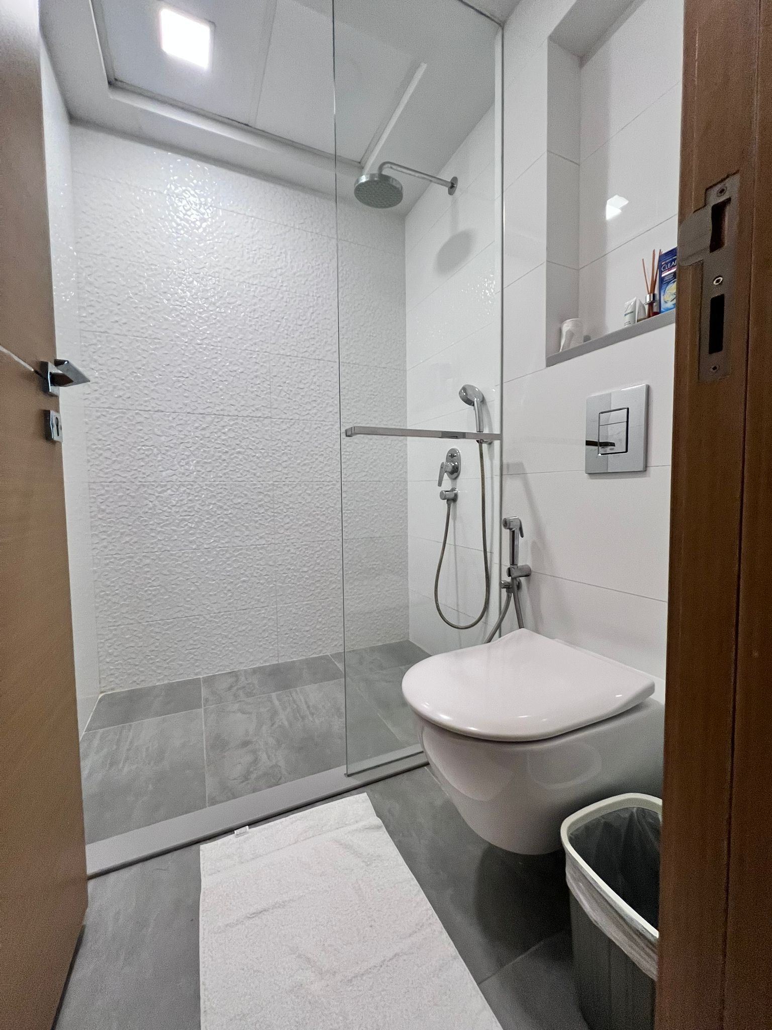 Modern bathroom with a walk-in shower, rain showerhead, and a wall-mounted toilet. Gray floor tiles and white walls. A white towel and trash bin are near the shower.