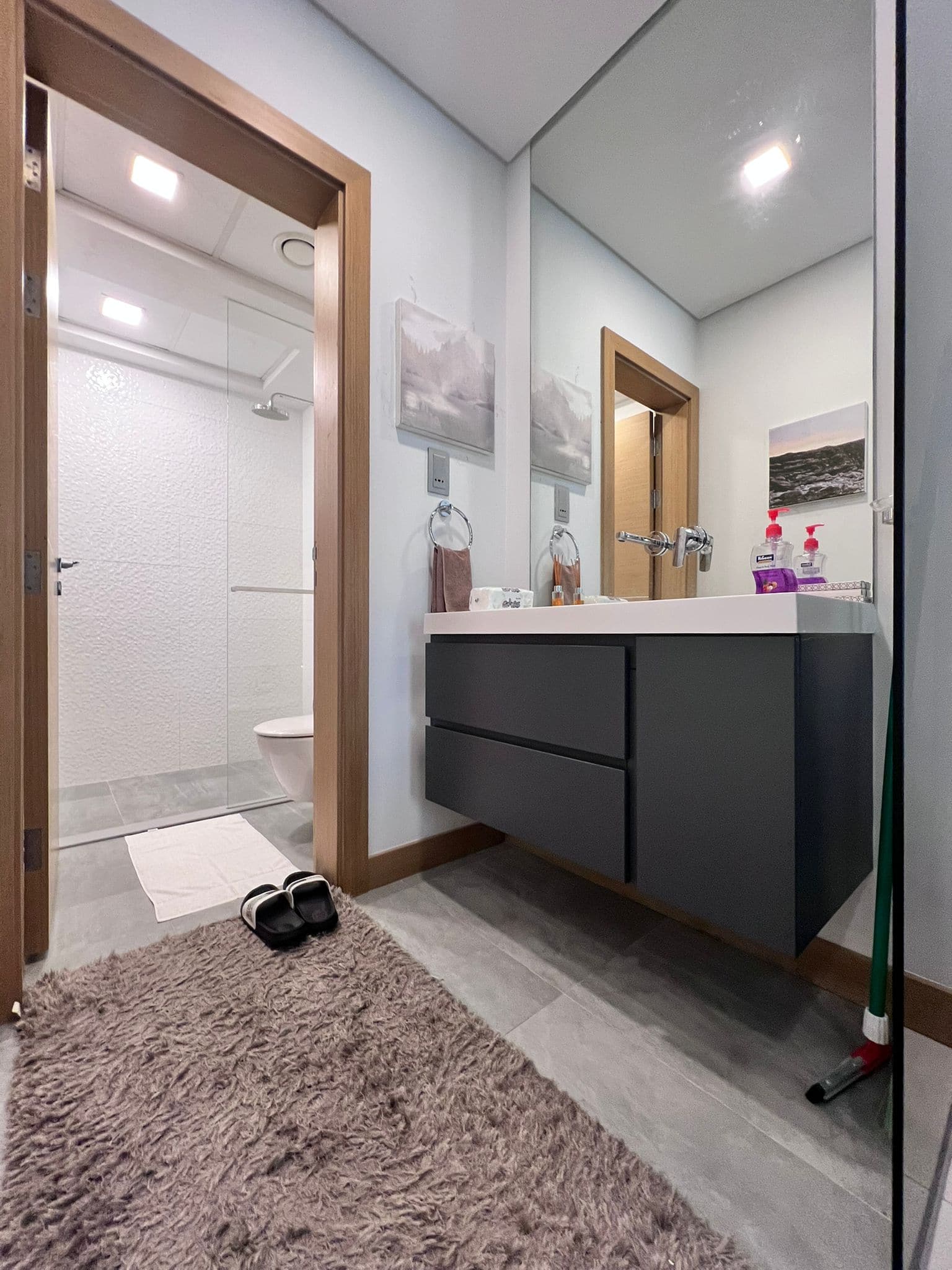 Modern bathroom with floating dark vanity, white sink, large mirror, and glass-enclosed shower. Gray rug on floor and slippers placed near doorway.