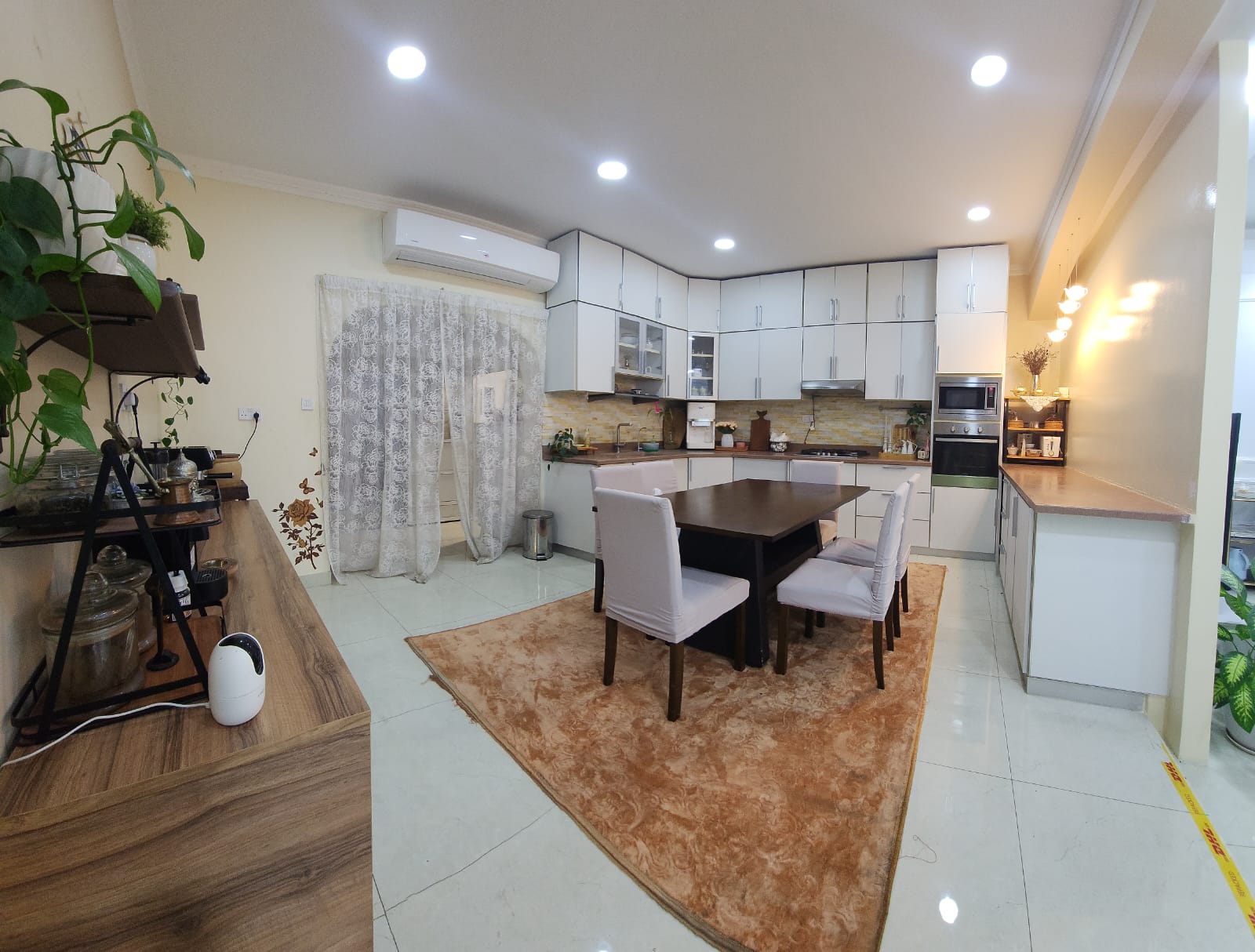 Modern kitchen with white cabinets, dining table with four chairs, brown rug, and plants. A curtain covers a doorway, and the room is well-lit with ceiling lights.