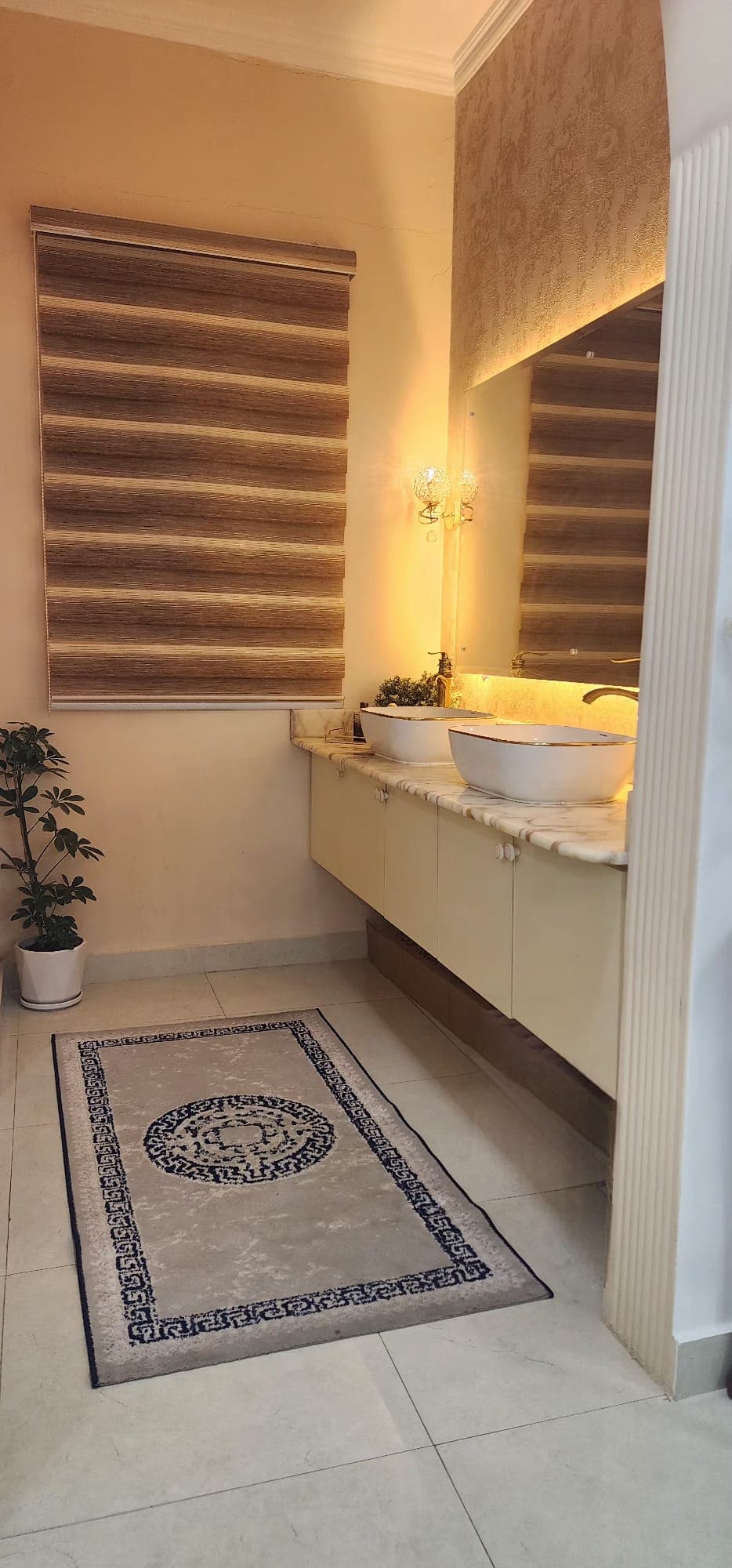 Modern bathroom with a large mirror, vessel sink on a marble countertop, potted plant, and a decorative rug on tiled floor. A window with a striped blind is on the left.