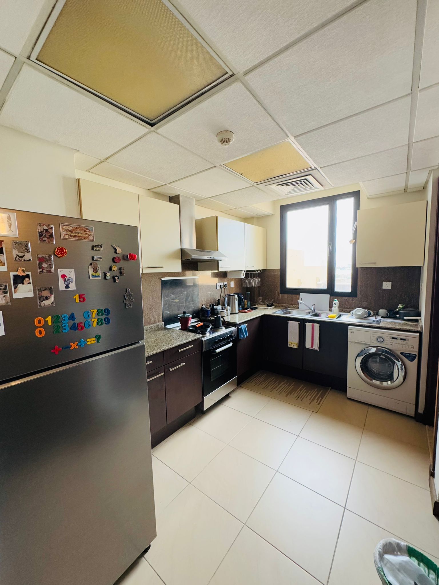 A kitchen with dark cabinets and beige countertops. Includes a fridge with magnets, oven, stove, microwave, sink, and washing machine. A window provides natural light.