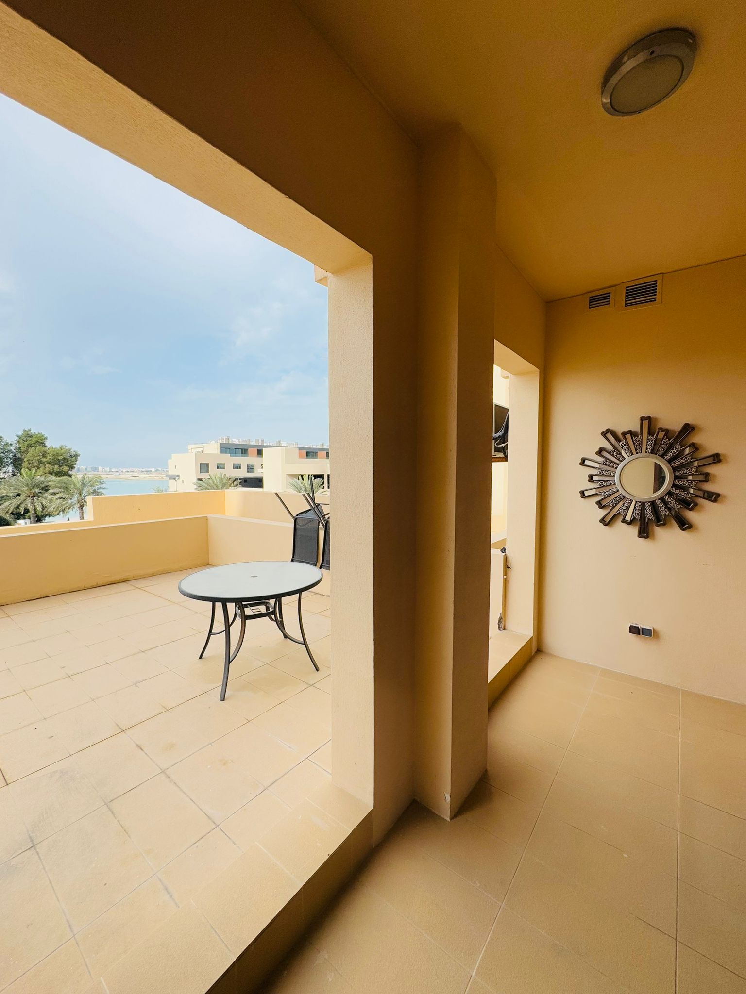 Covered balcony with beige walls, a round mirror on the wall, and a round table with two chairs. The balcony overlooks trees and buildings under a cloudy sky.