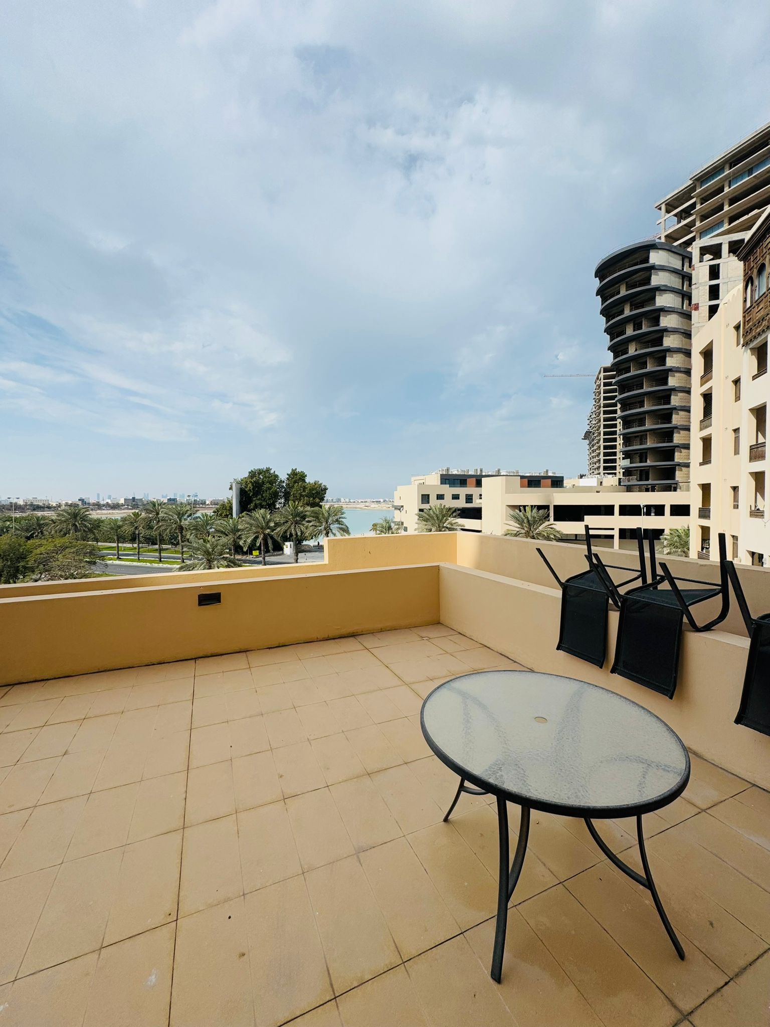 Balcony with a round glass table and four inverted chairs. Overlooking a view of palm trees, buildings, and a distant body of water under a cloudy sky.