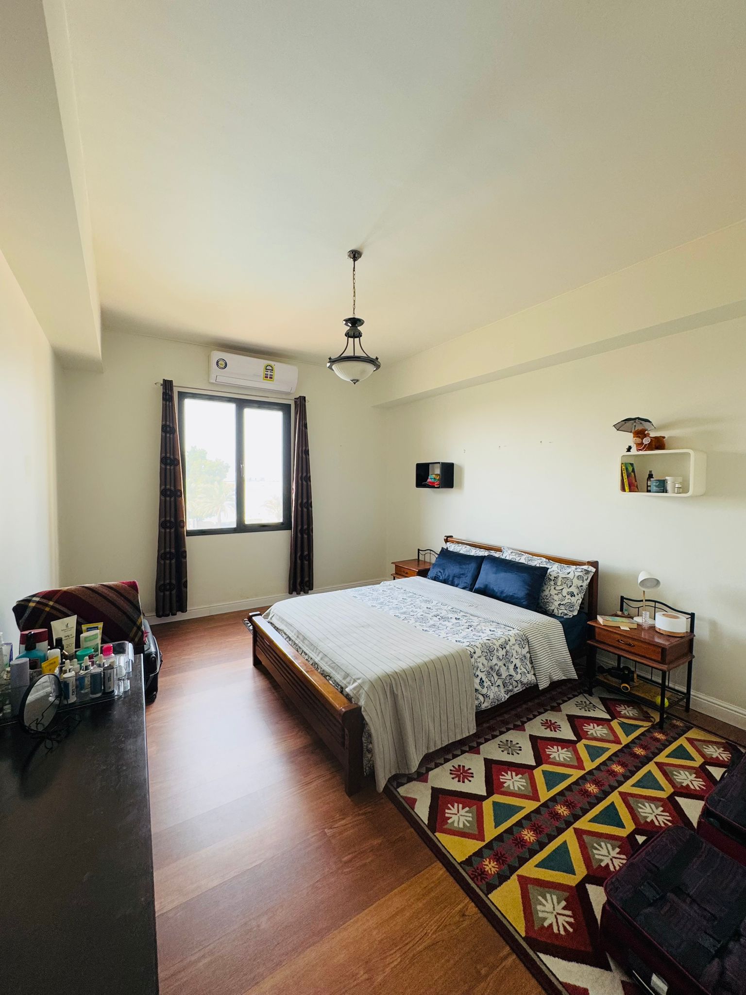 A tidy bedroom with a window. It has a bed with a blue and white bedspread, a side table with a lamp, a dresser with toiletries, shelves, and a colorful rug on wooden flooring.