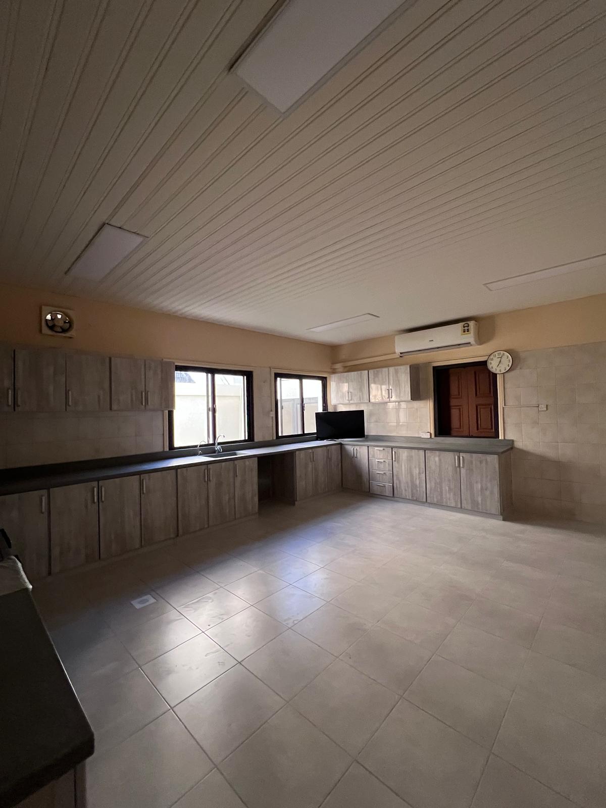 Spacious kitchen with tiled floor, wooden cabinets, black countertops, and a wall-mounted clock. Ceiling has recessed lighting. Air conditioning unit above the door. Large windows provide natural light.