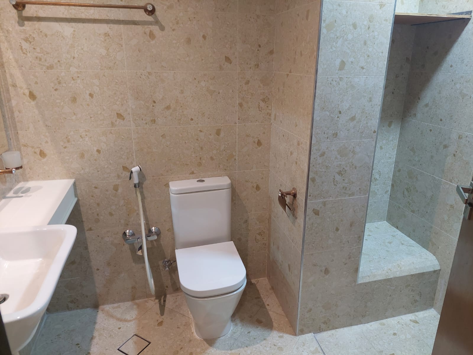 Bathroom with beige tiled walls, featuring a white toilet, a wall-mounted sink to the left, and an open shower area to the right. A toilet spray is positioned next to the toilet.