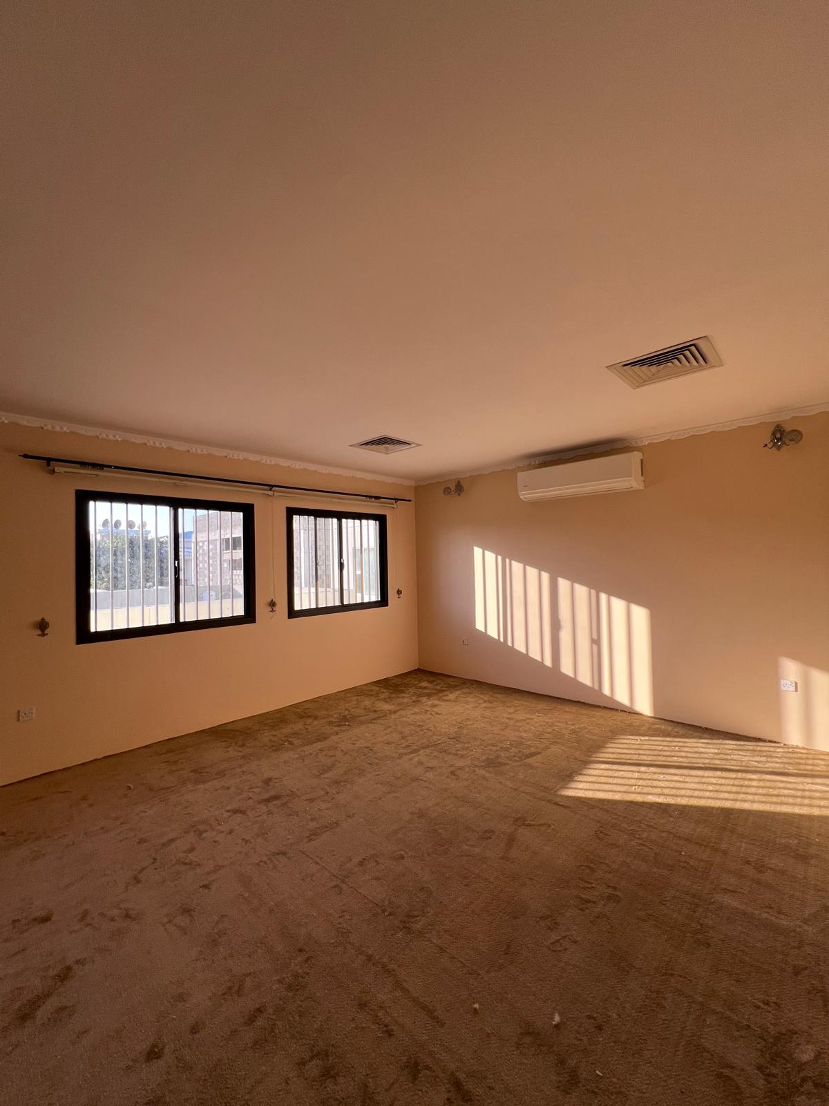 Empty room with beige walls and carpet, three windows with bars, air conditioning unit, and ceiling lights. Sunlight casts shadows on the floor.