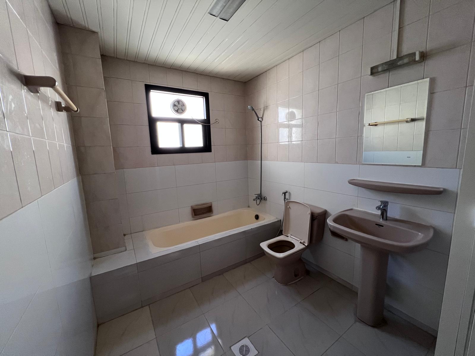 Bathroom with beige fixtures, including a bathtub, toilet, and sink. Tiled walls and floor, a window above the tub, a mirror above the sink, and a showerhead are visible.