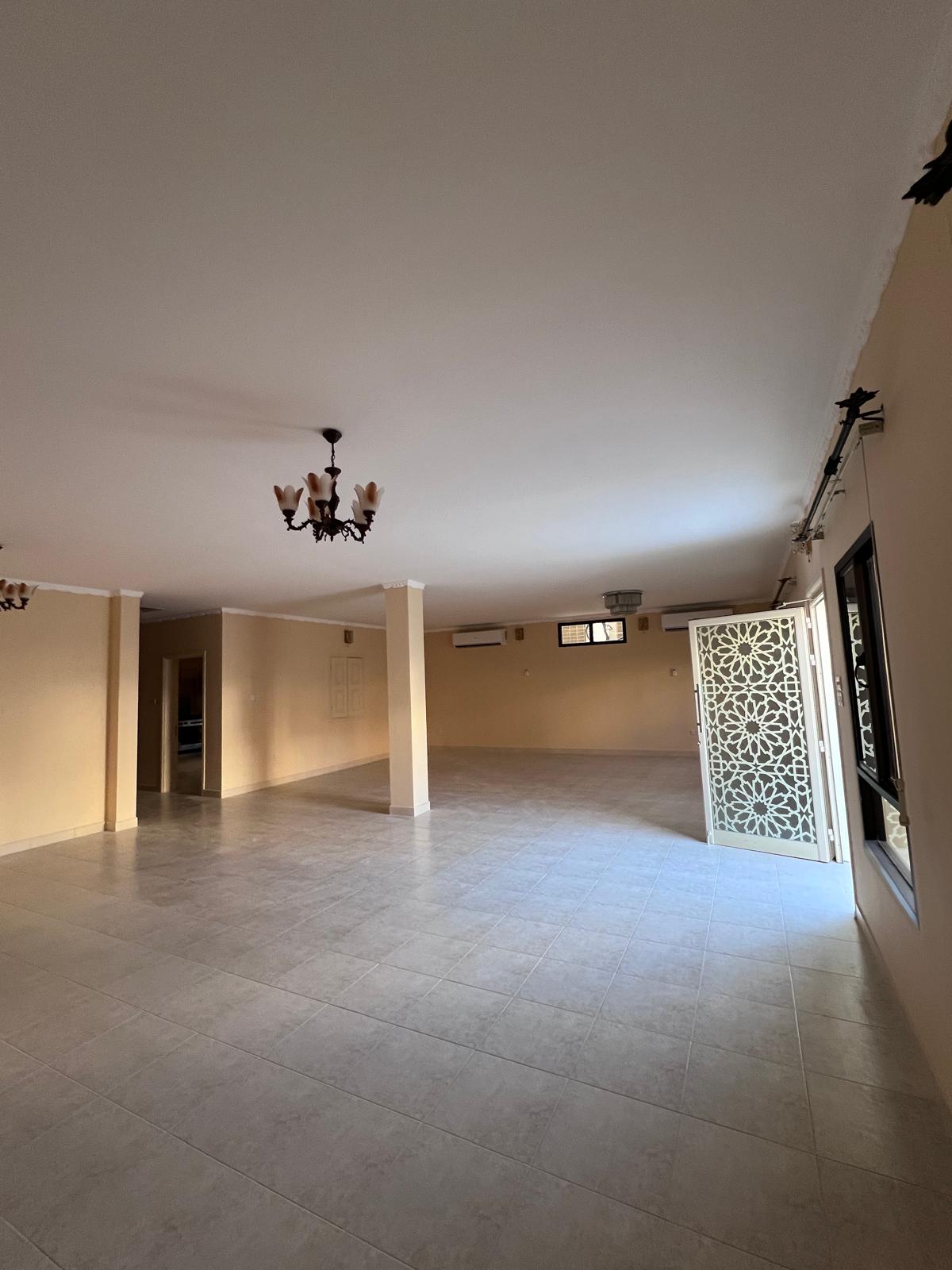 Spacious empty room with beige walls, tiled floor, ornate white door, and ceiling lights. There are small windows and an air conditioning unit on the far wall.