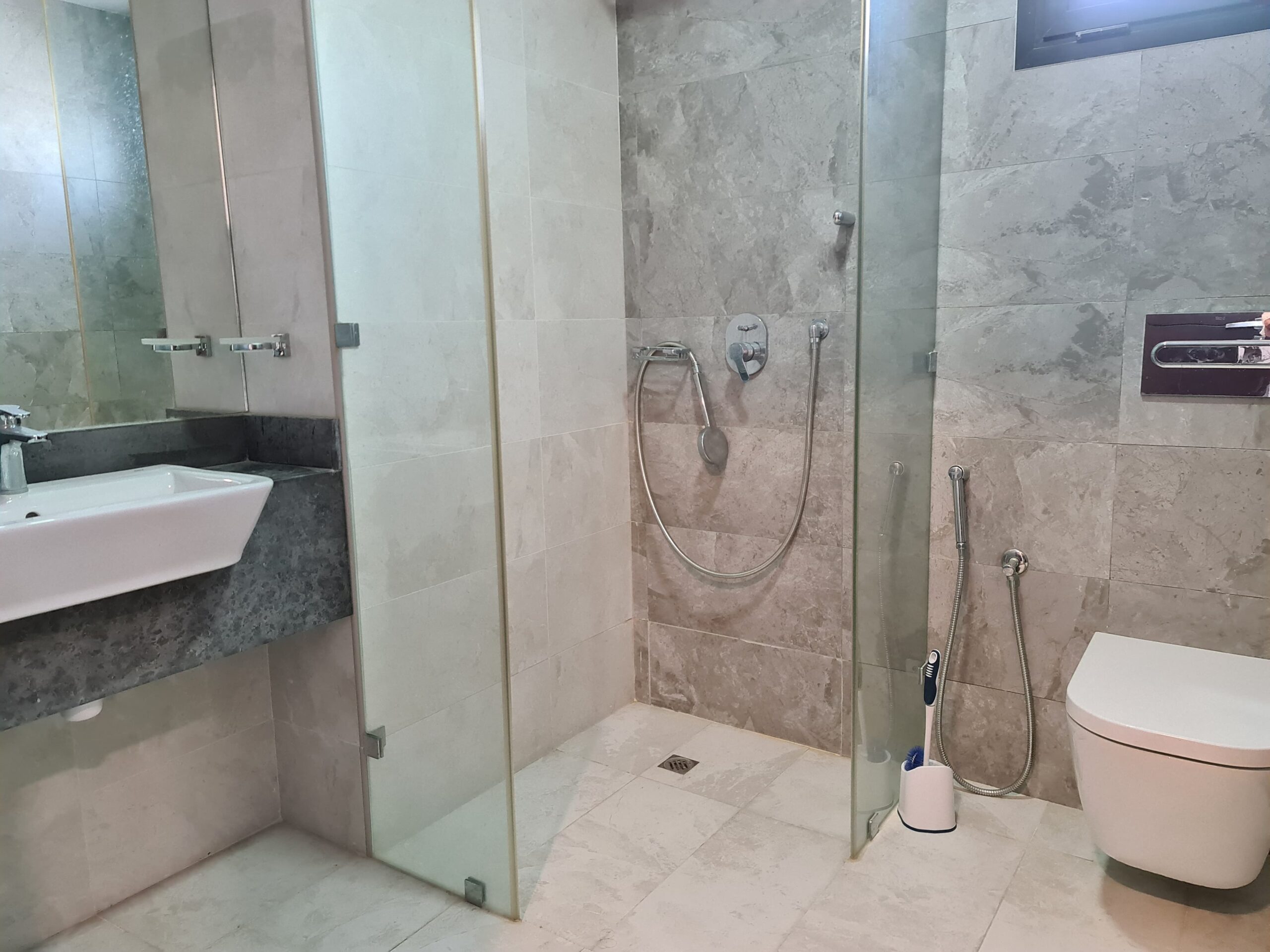 Modern bathroom with a glass-enclosed shower, wall-mounted toilet, sink with countertop, and gray stone tiles. Showerhead and faucet are visible. Toilet brush and spray hose present.