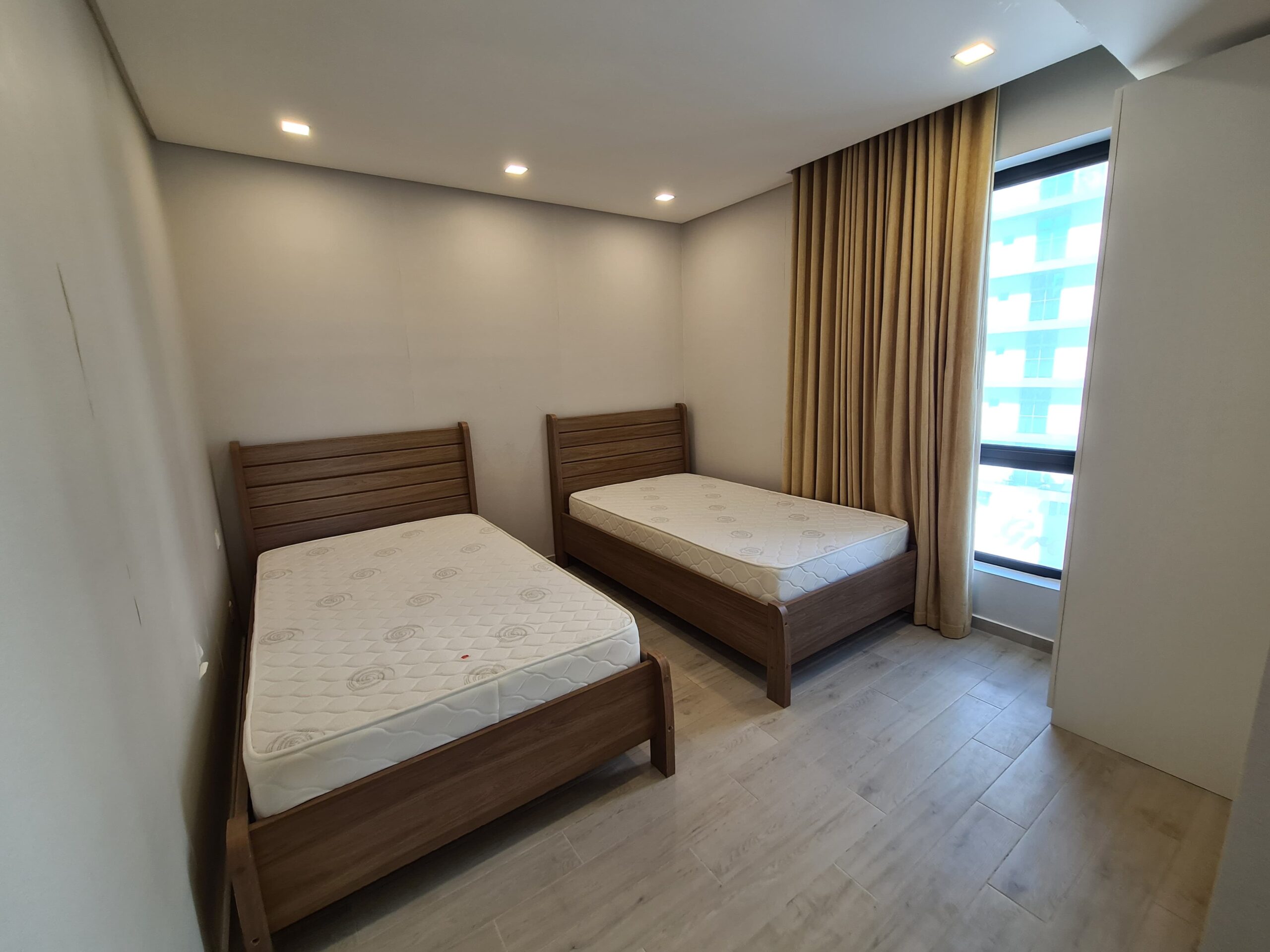 Room with two wooden single beds, mattresses, beige curtains, and a large window. Walls and floor are light-colored. Ceiling lights illuminate the space.