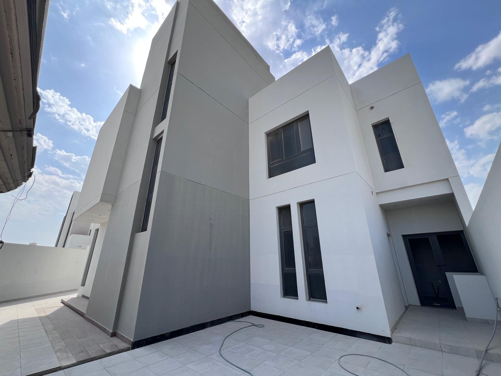 Modern white and gray building with geometric design, large windows, and a tiled patio under a partly cloudy blue sky.