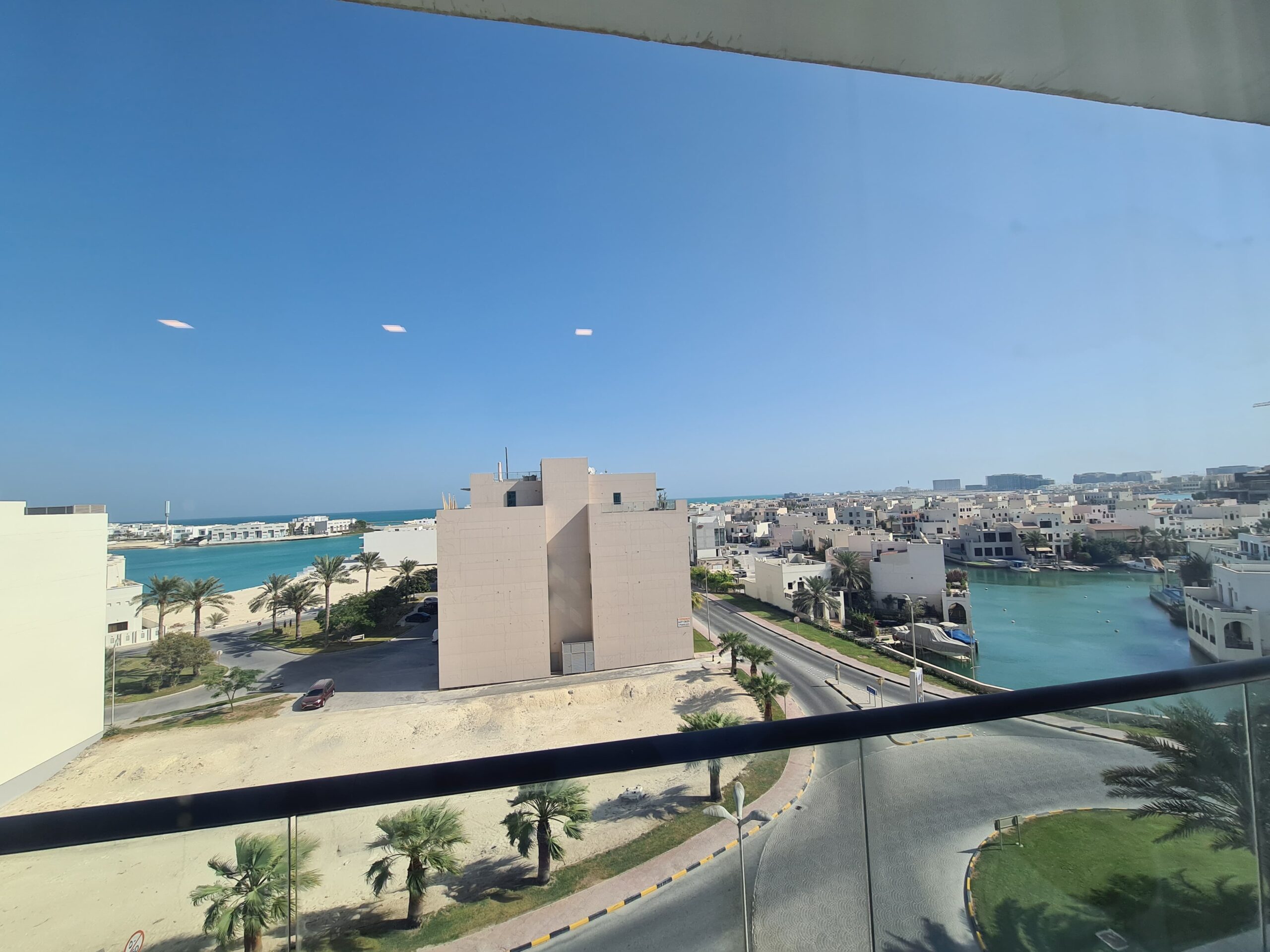 View from a balcony shows modern beige buildings, a road with palm trees, and a waterway under a clear blue sky.