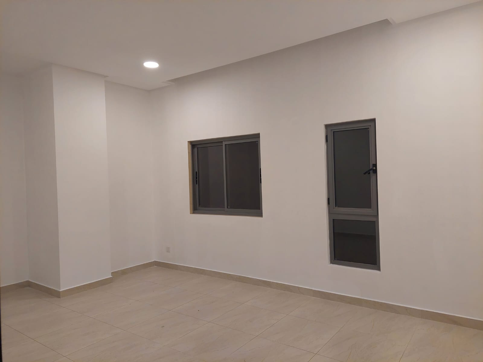 Empty room with beige tiled floor, white walls, a small window and a narrow vertical window. Ceiling light is on.