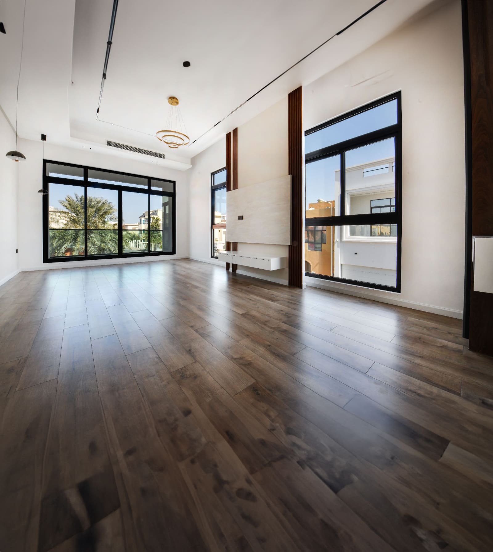 Spacious empty room with wooden flooring, large windows, a wall-mounted shelf, and a modern light fixture on the ceiling. View of trees and buildings outside.