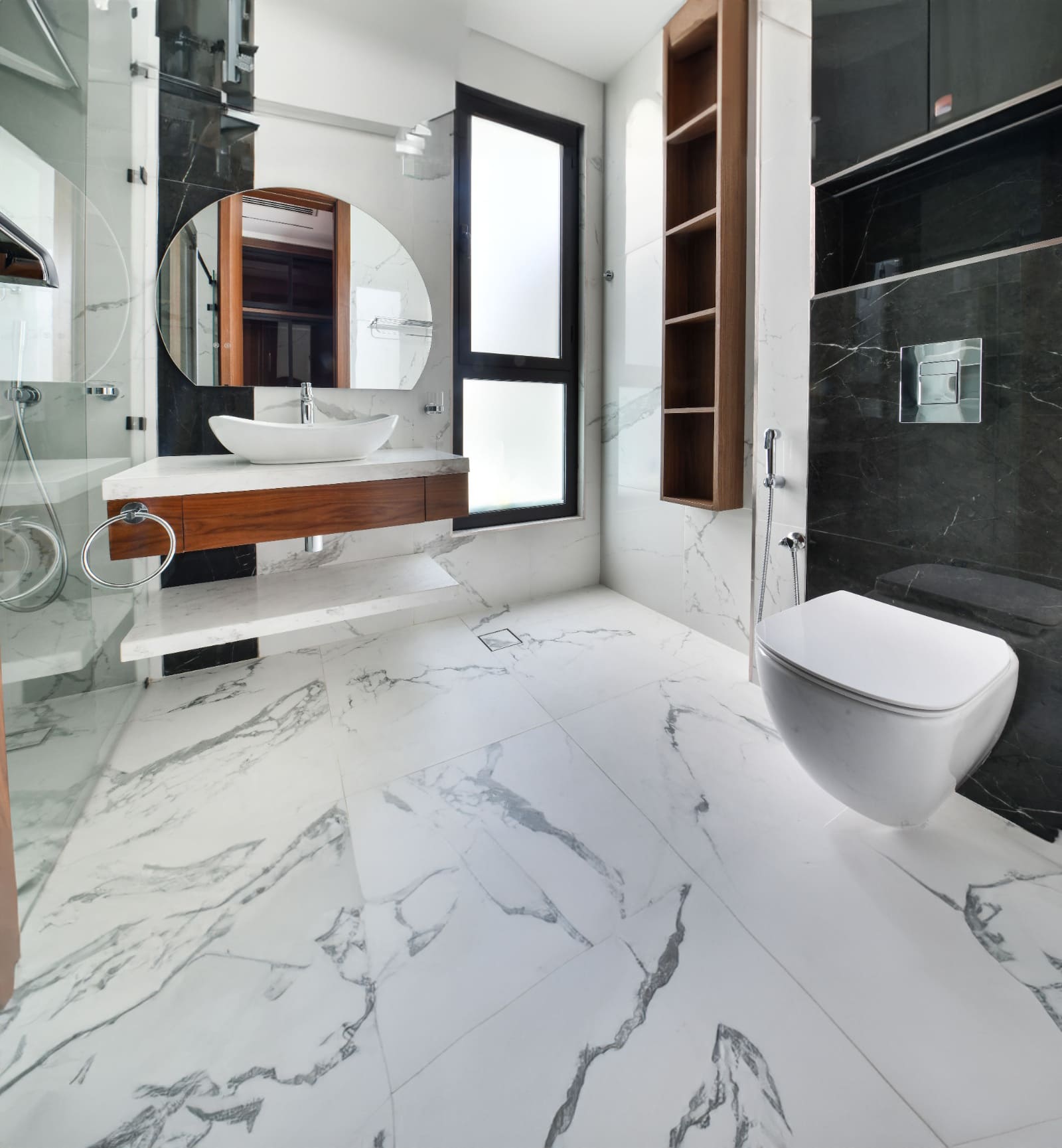 Modern bathroom with marble flooring, circular mirror, white sink on a wooden counter, and a wall-mounted toilet beside large frosted glass windows.