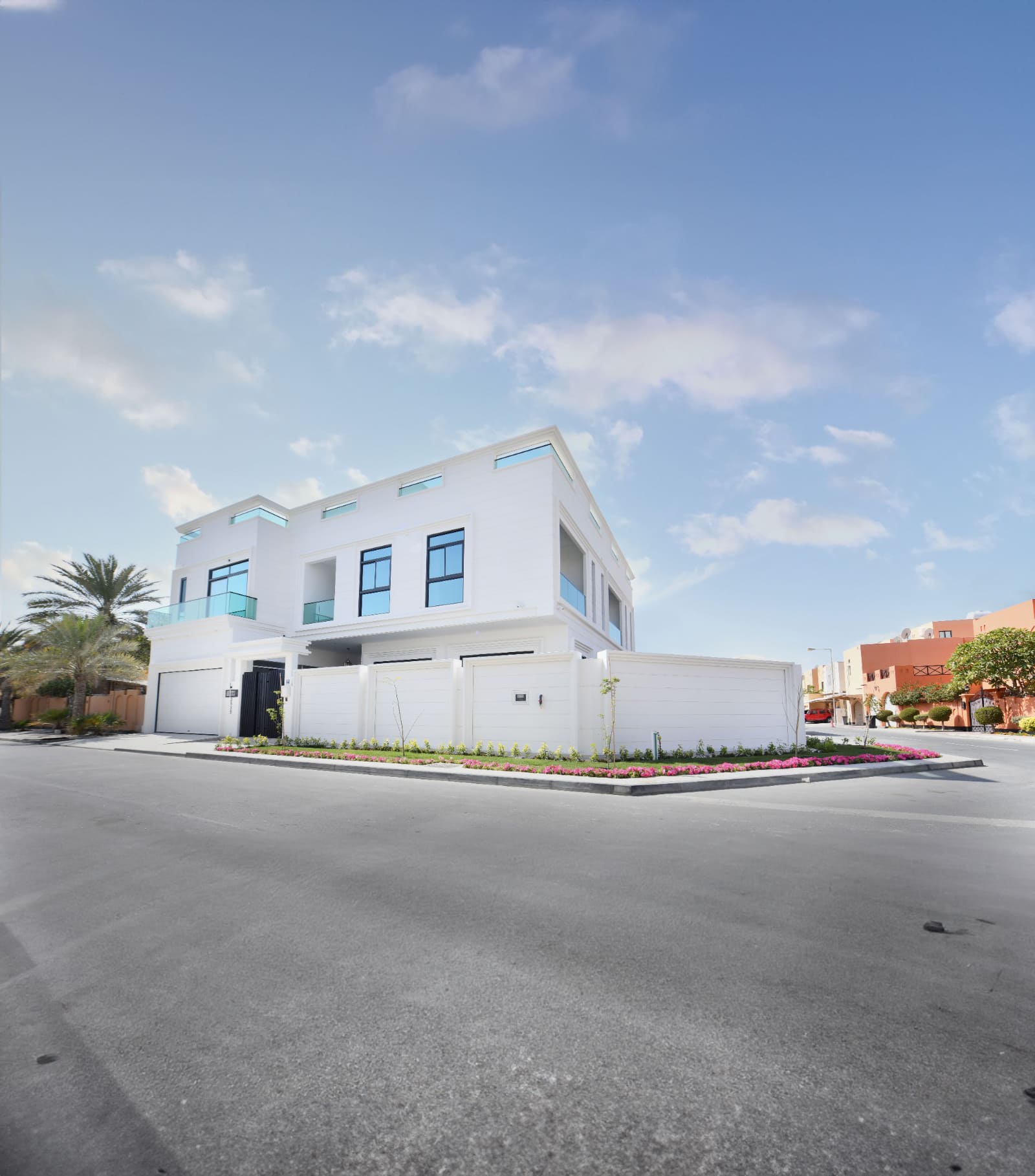 Modern white two-story house with large windows on a corner lot, surrounded by a landscaped garden. Clear blue sky and neighboring buildings in the background.