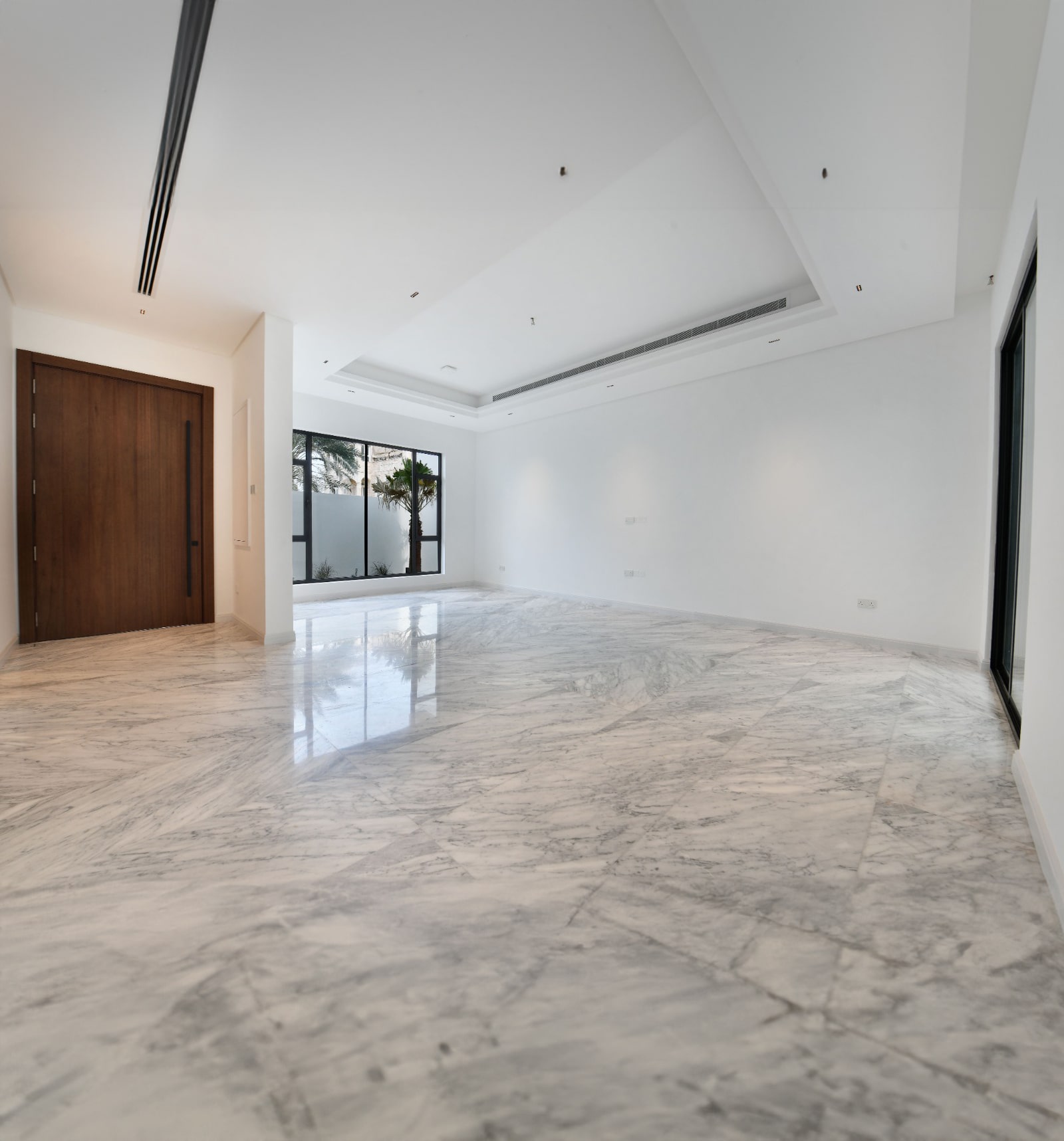 Spacious empty room with marble flooring, white walls, a large wooden door, and windows offering natural light.