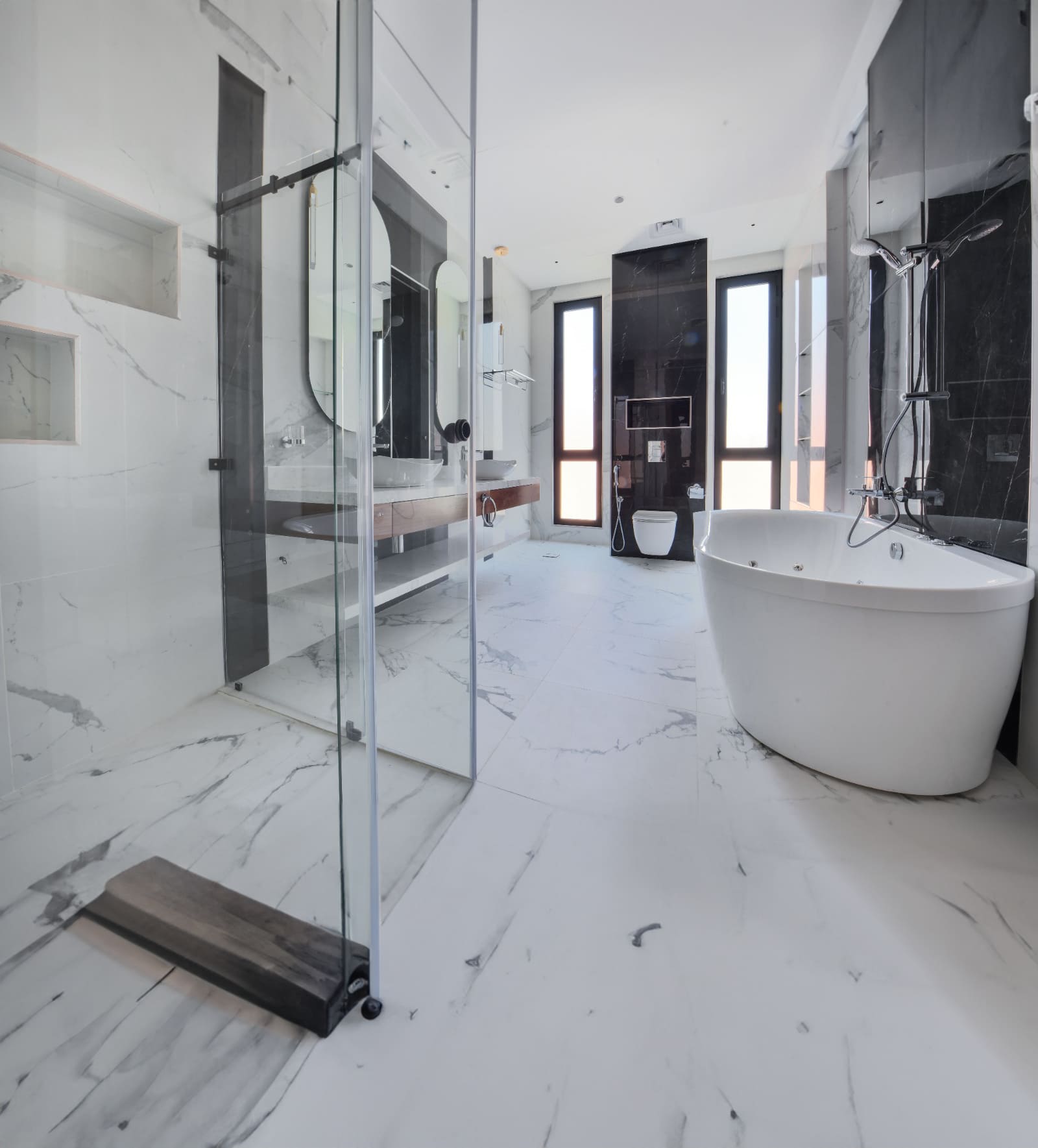 Modern bathroom with white marble flooring, a glass-enclosed shower, freestanding bathtub, dual sinks, and two tall windows.