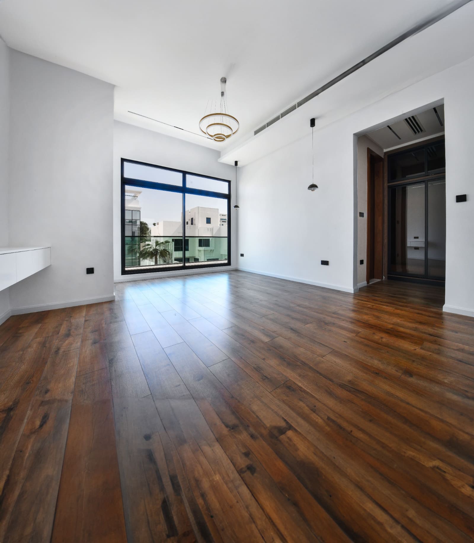 Spacious empty room with wooden floor, large window, white walls, and a modern chandelier.