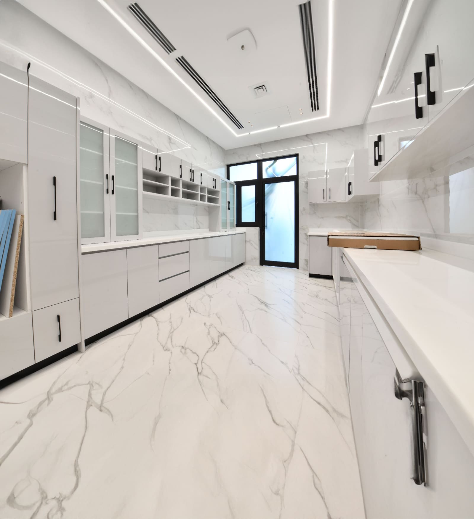 A modern, sleek kitchen with white marble floors and countertops, glass cabinet doors, and stainless steel handles. Bright lighting enhances the spacious, contemporary design.