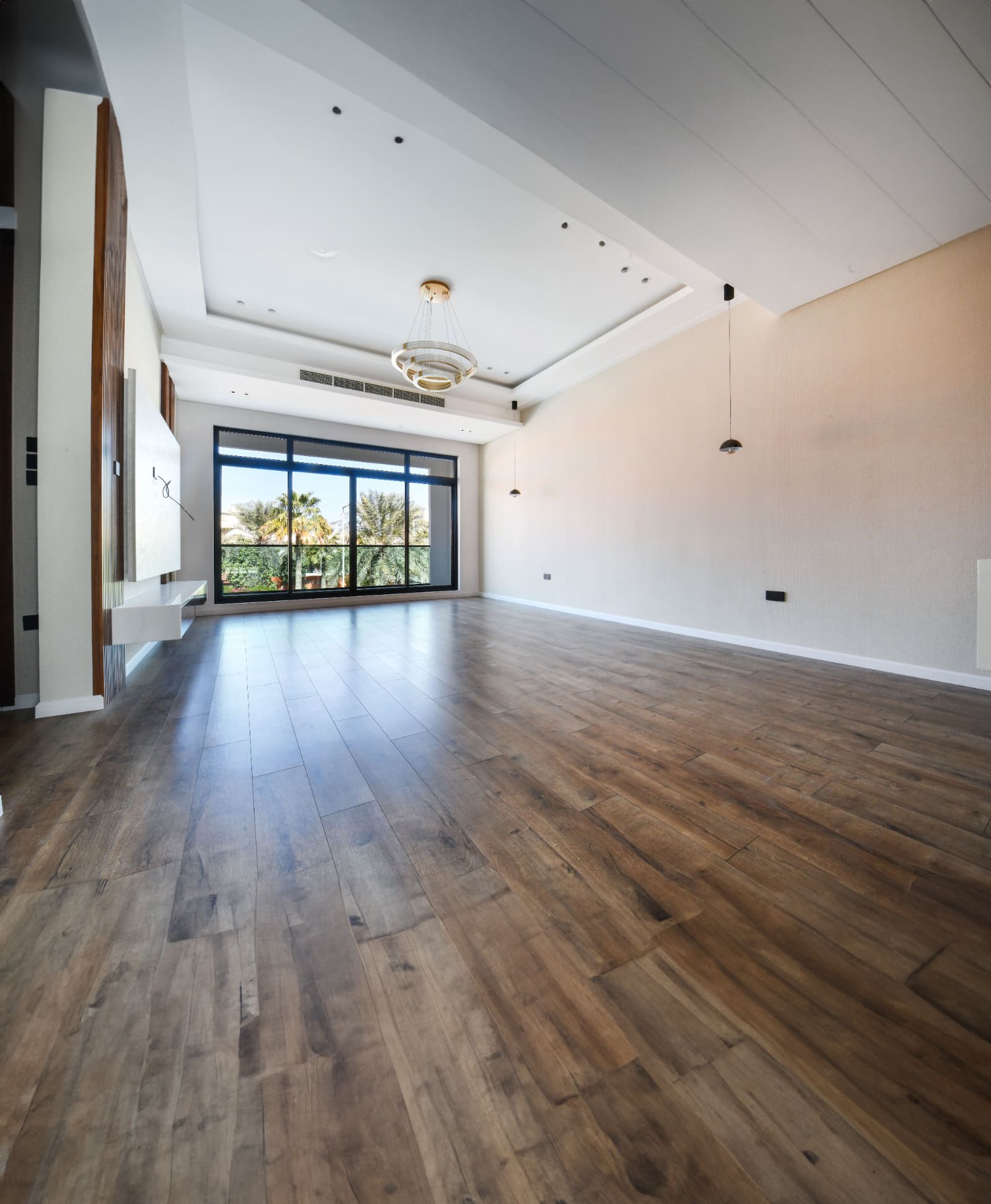 A spacious, empty room with wooden flooring, large windows, pendant lighting, and a chandelier. The windows overlook a view with palm trees.