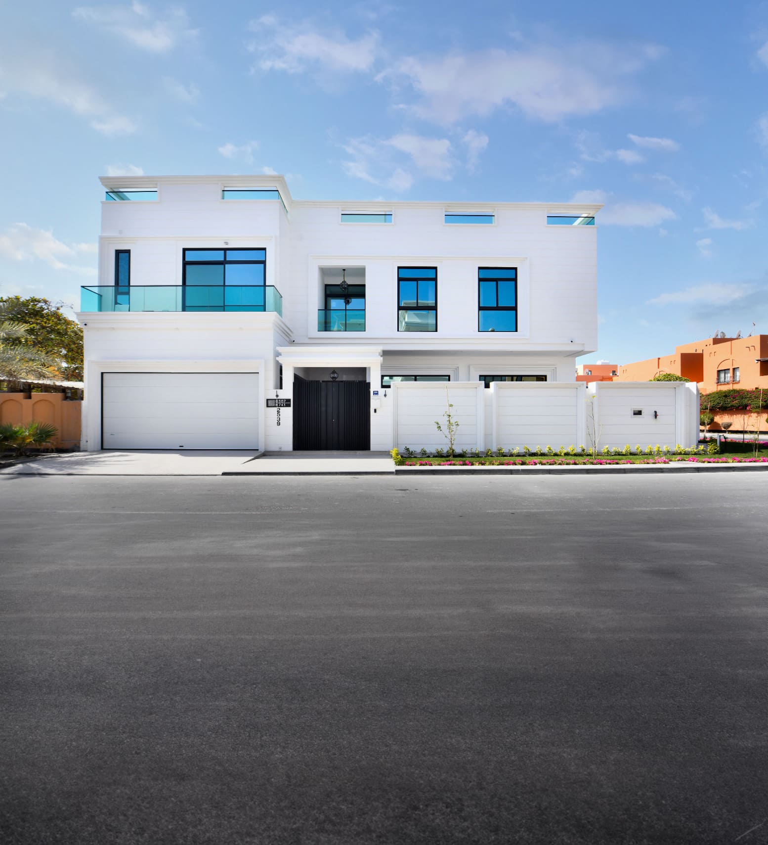 Modern white two-story house with large windows, a garage, and a balcony. The street in front is empty, and the sky is clear with a few clouds.