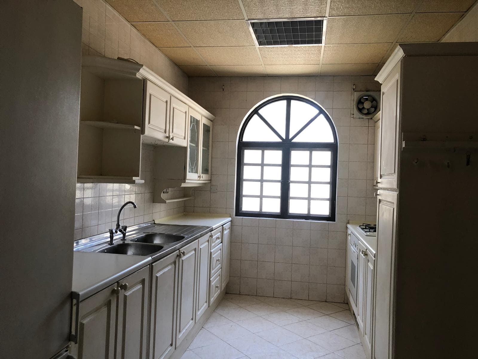 A kitchen with white cabinets, tiled walls, a double sink, gas stove, oven, and an arched window with a black frame, allowing natural light to fill the space.