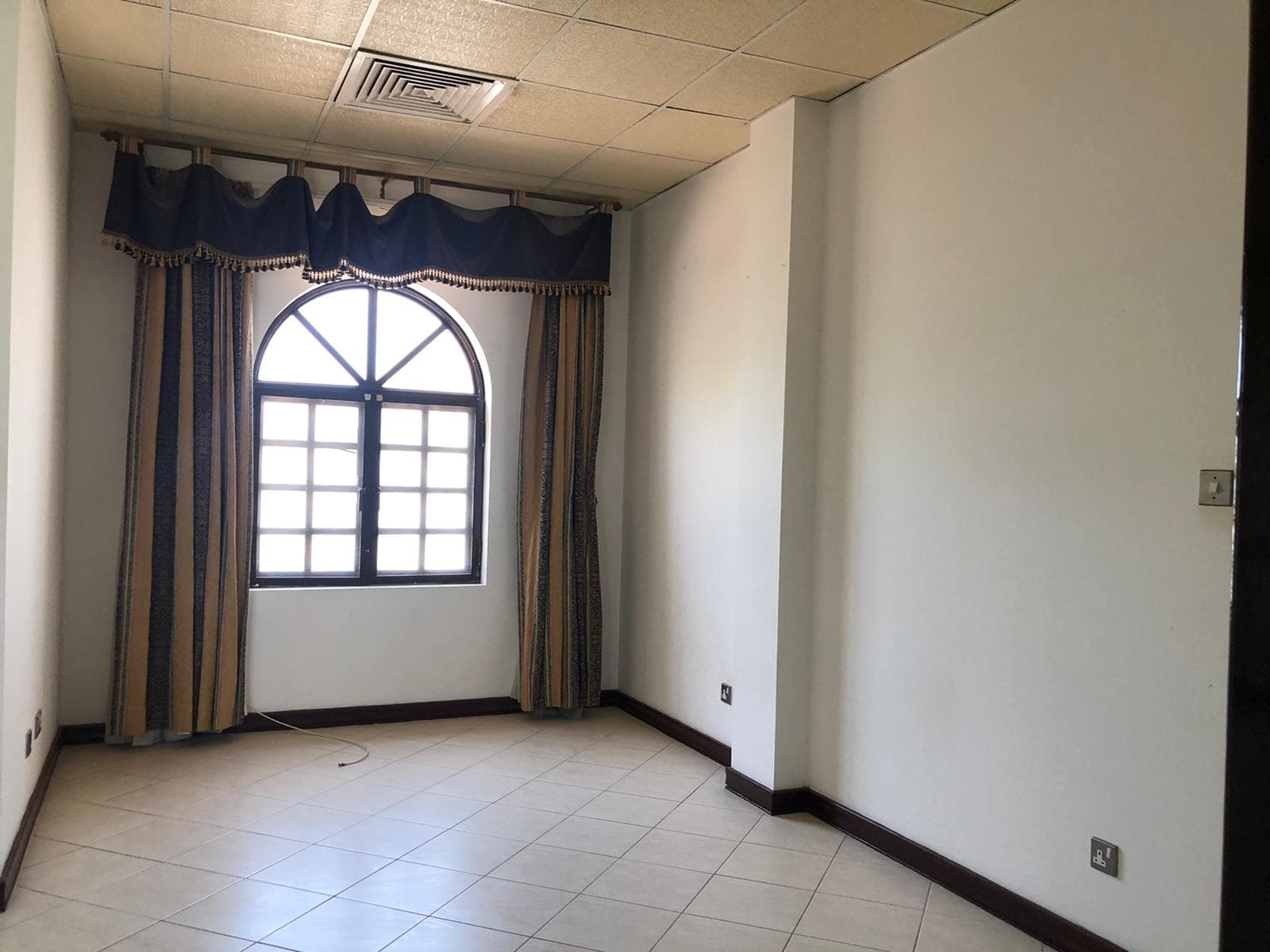 A small, empty room with tiled flooring, a large arched window with blue and gold curtains, and a ceiling with recessed lighting. The walls are plain and painted off-white.