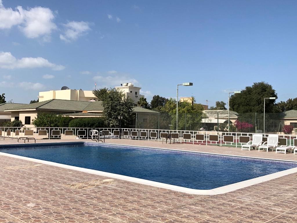 An outdoor swimming pool surrounded by a tiled deck with lounge chairs and greenery in the background.