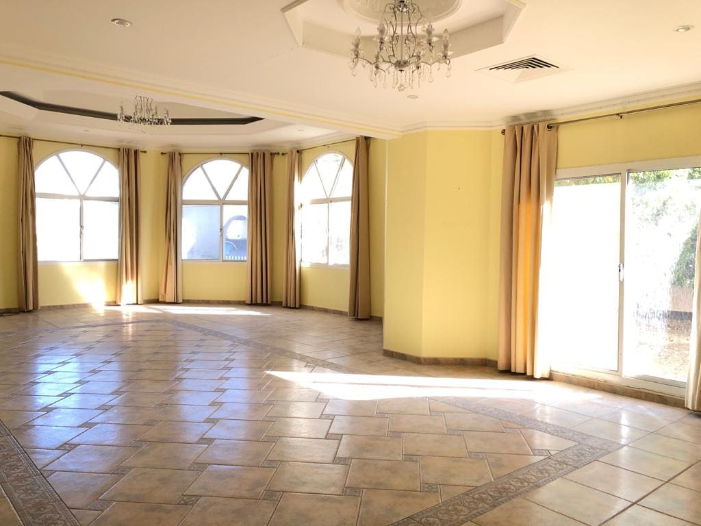 An empty, sunlit room with tiled flooring, multiple arched windows with beige curtains, a sliding glass door, and a ceiling chandelier.