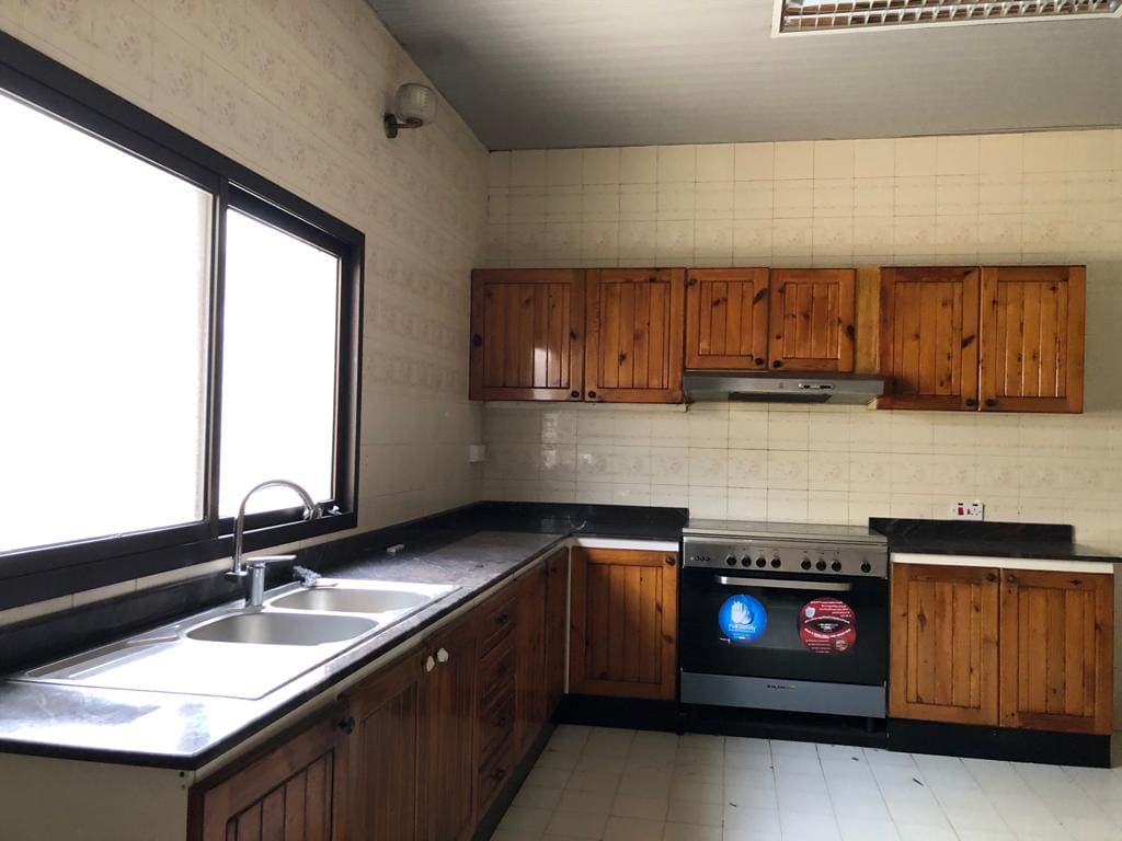 A kitchen with wooden cabinets, a double sink on the left counter, a stove with an oven in the center, and a large window above the sink. The walls and floor are tiled.