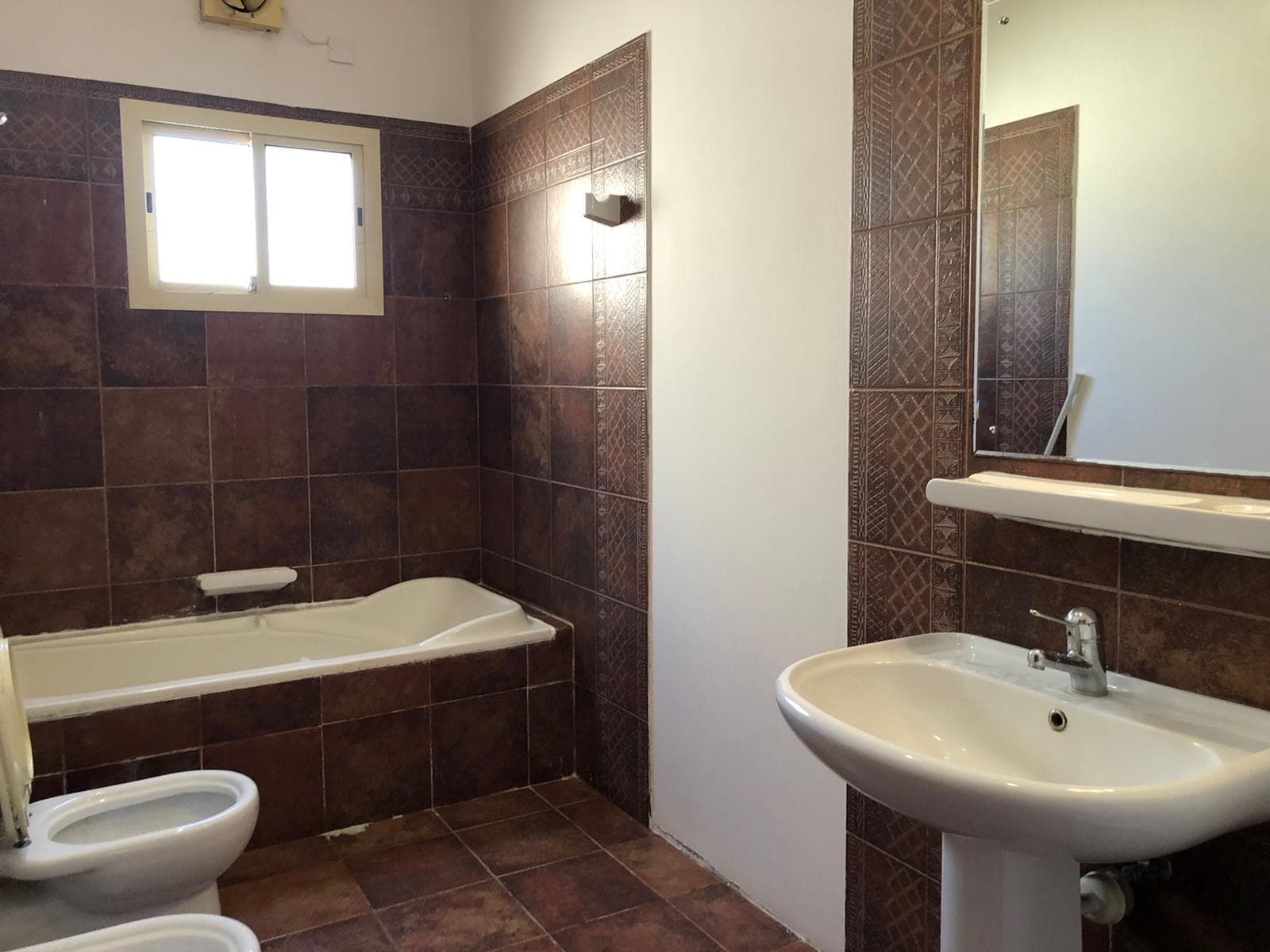 A bathroom with a bathtub, bidet, toilet, and sink. The walls and floor are tiled in dark brown, and there is a window above the bathtub. A mirror is mounted above the sink.